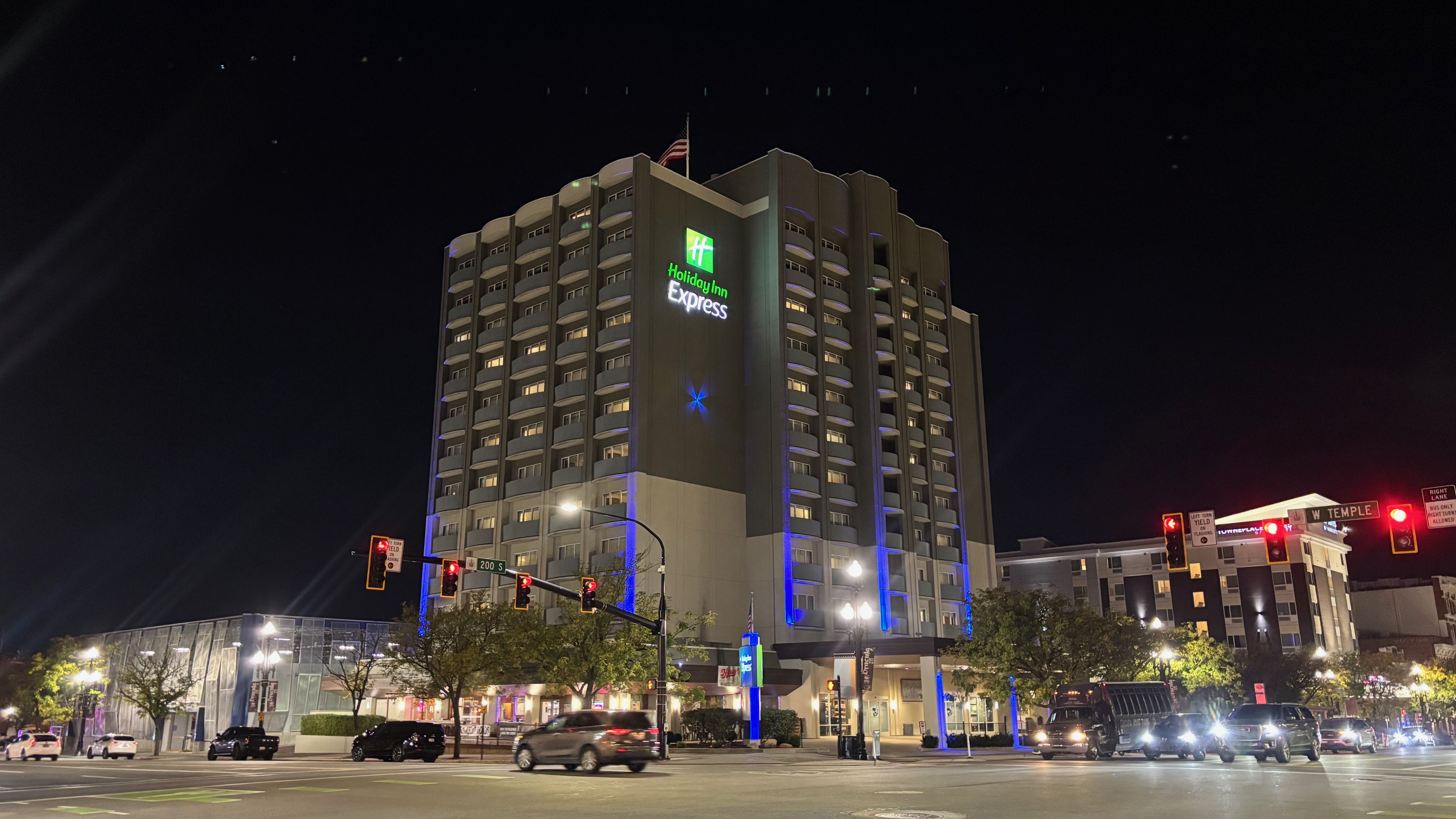Night view of a tall Holiday Inn Express with curved balconies, green logo, American flag, and blue accent lighting at a busy intersection with red traffic lights.
