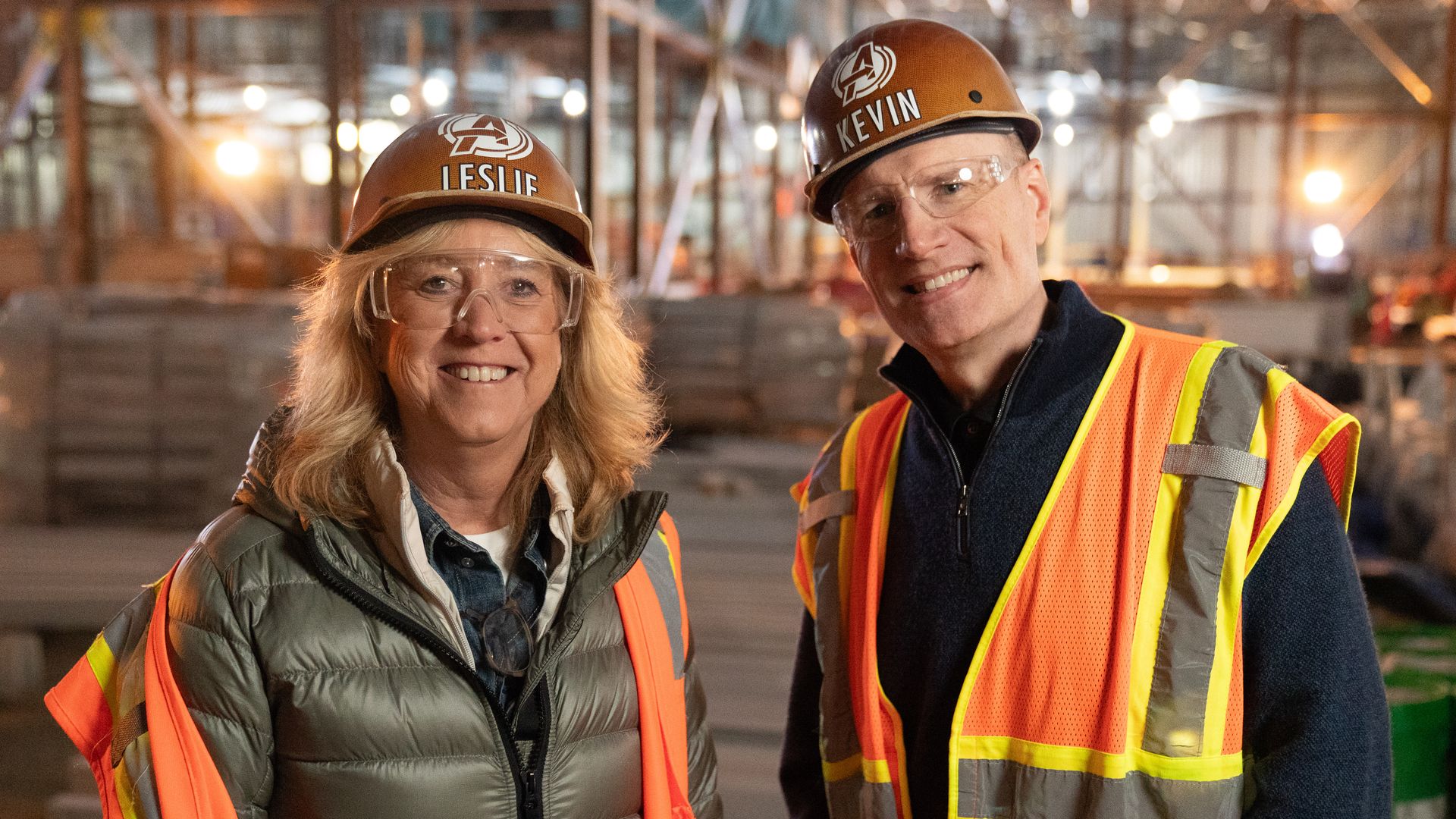 Two smiling construction workers wearing brown hard hats labeled LESLIE and KEVIN, clear safety goggles, and bright orange safety vests, posing at a construction site with scaffolding in the background.