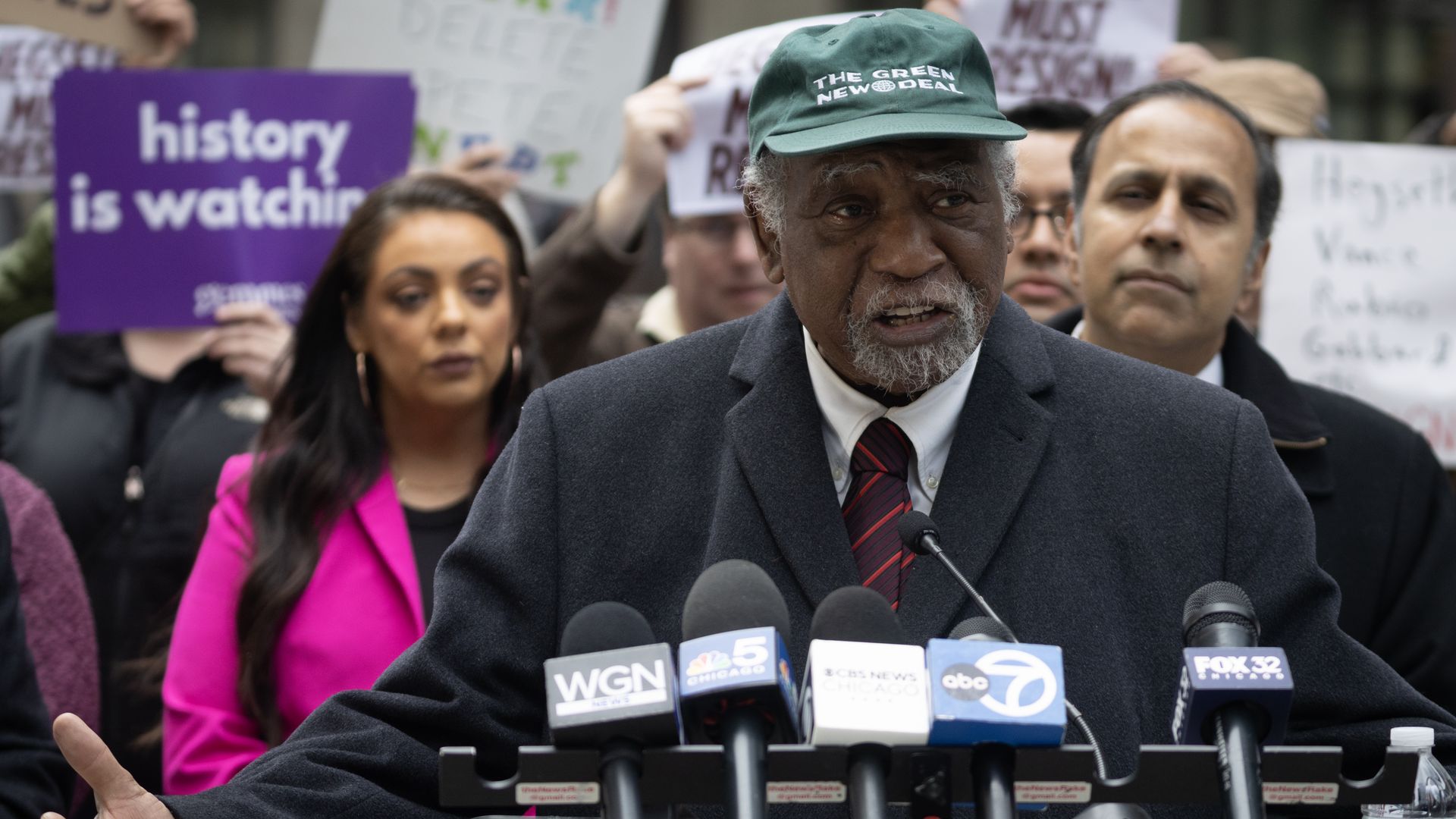 An older man with a gray beard in a green "The Green New Deal" cap and dark coat speaks at a podium with multiple news microphones, while people stand behind him holding protest signs.