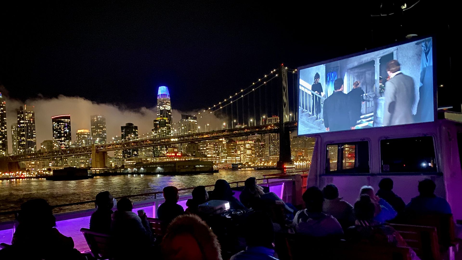 People watching a movie on an outdoor screen at night on a boat with purple lighting, city skyline and illuminated bridge in the background over water.