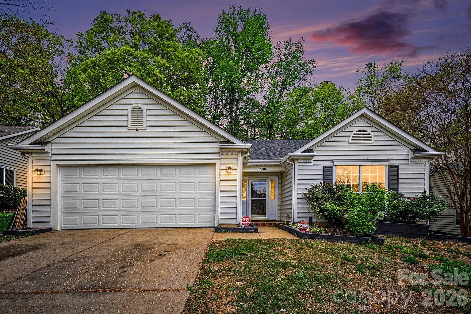 White, single-story house with two front gables and a two-car garage on the left. Center-front door, window with shutters on the right, shrubs in front, green trees behind, purple sunset sky.