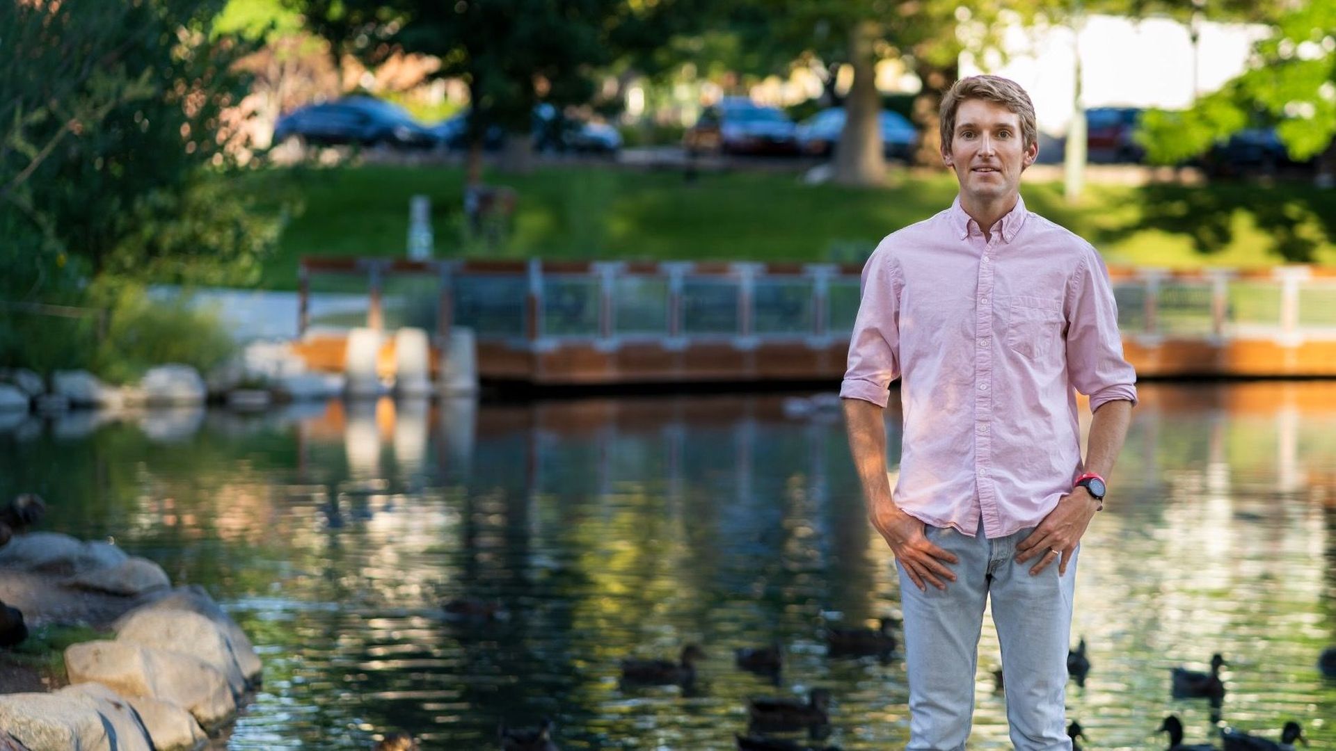 A man in a pink shirt and blue jeans stands in front of a park pond.