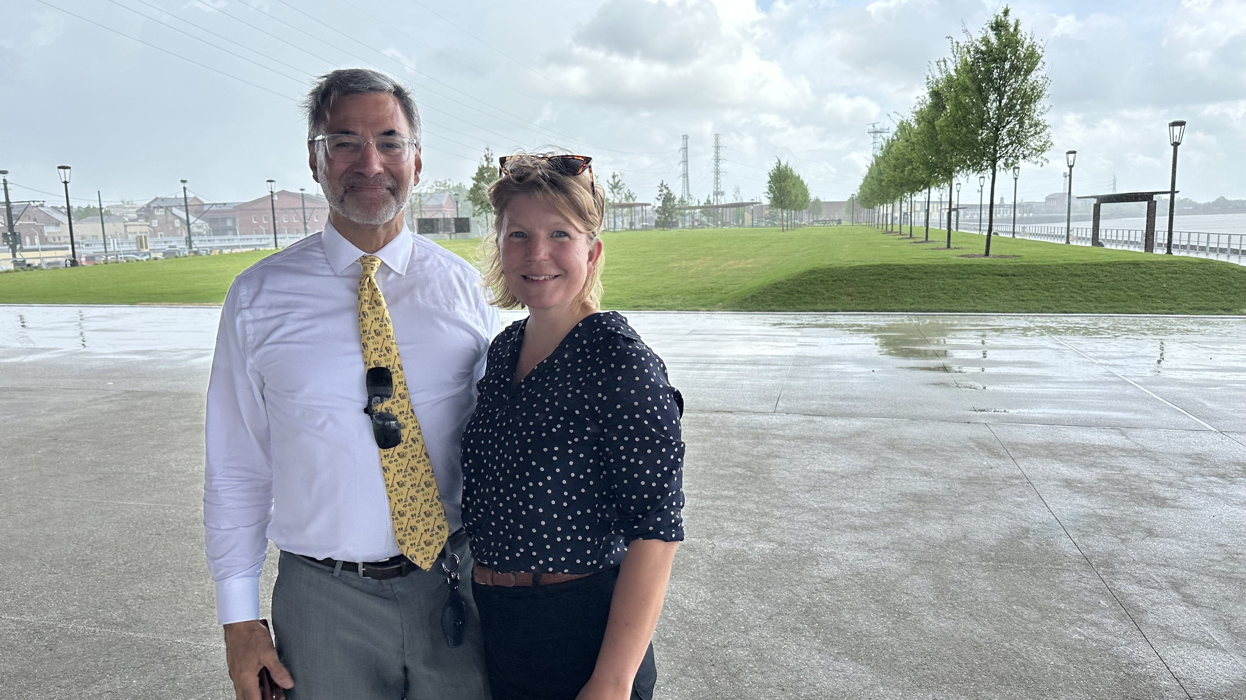 Two adults stand outdoors on a rainy day by waterfront park: man in a white shirt with a yellow tie and sunglasses, woman in a navy polka-dot blouse with a brown belt; wet pavement, green lawn, trees.