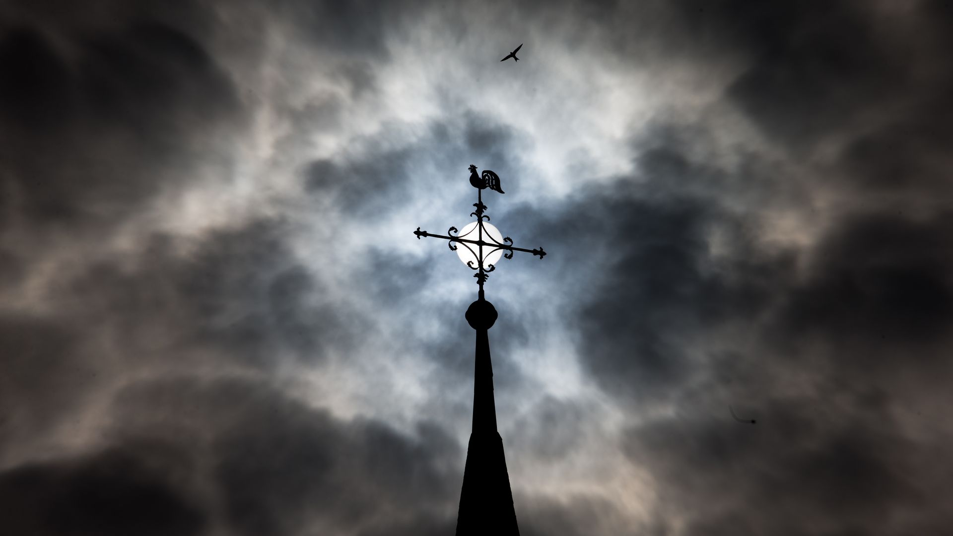 Weather cock and cross on top of a Catholic church in Germany, amid dark storm clouds.