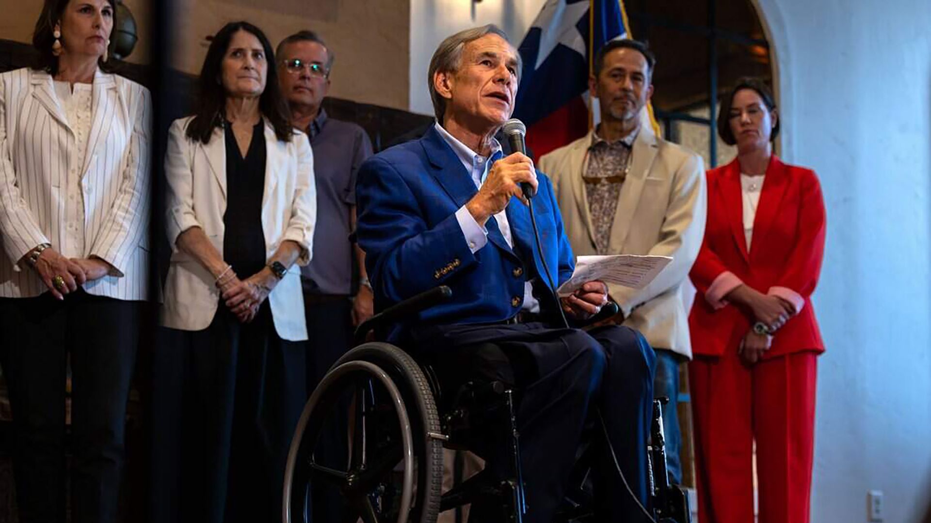 Texas Gov. Greg Abbott speaks into a microphone while seated in a wheelchair, with several people standing behind him.