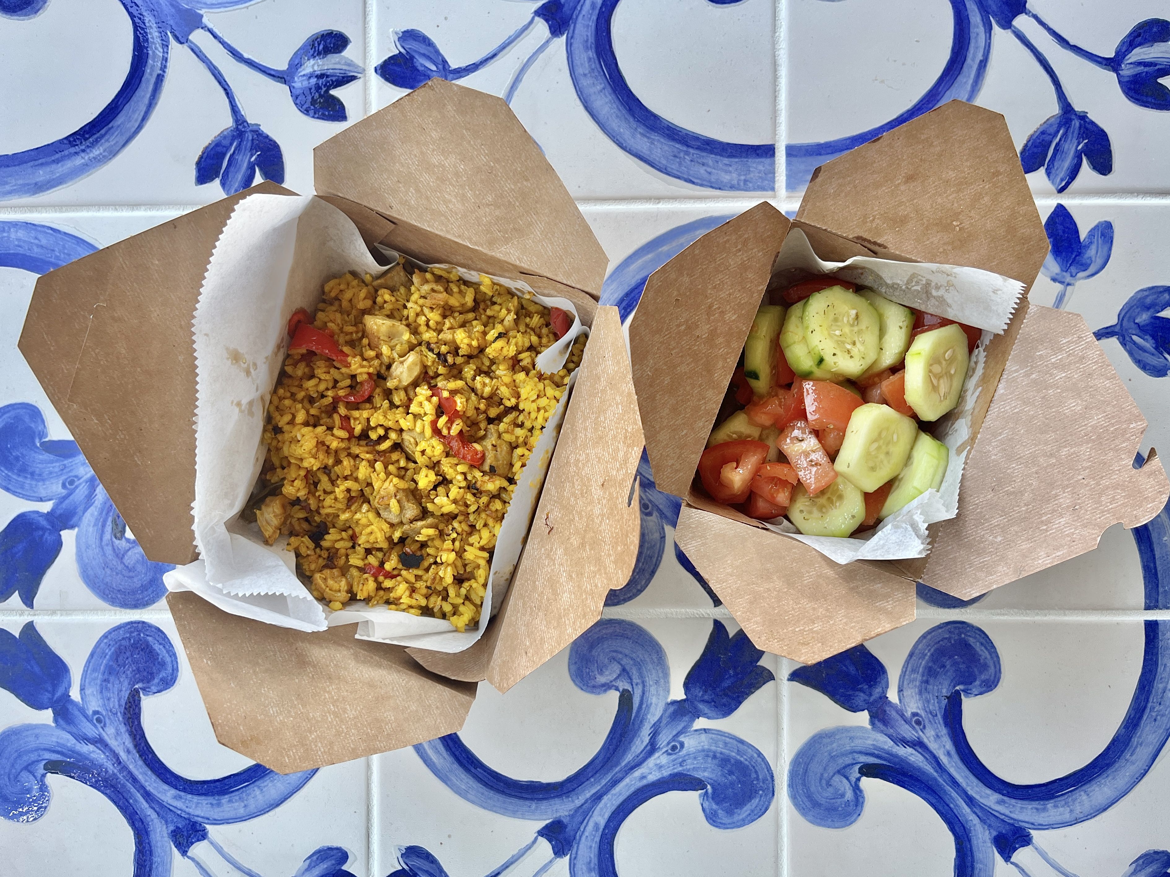 Two open brown takeout boxes on blue and white patterned tiles. One contains yellow rice with chicken and red peppers; the other has cucumber and tomato salad with seasoning.