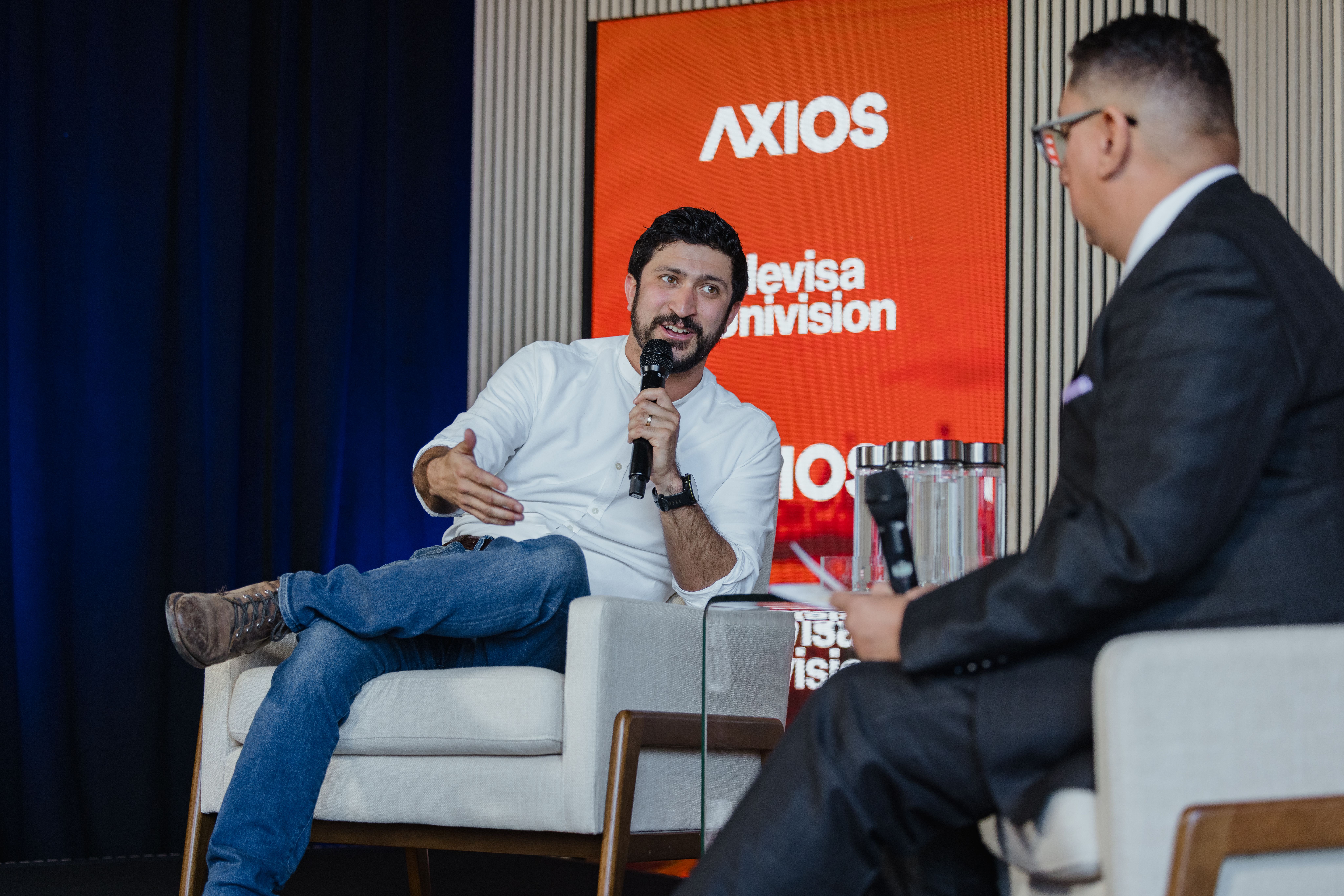Two men on a stage; one in a white shirt and jeans speaks into a microphone and gestures, while the other in a dark suit listens