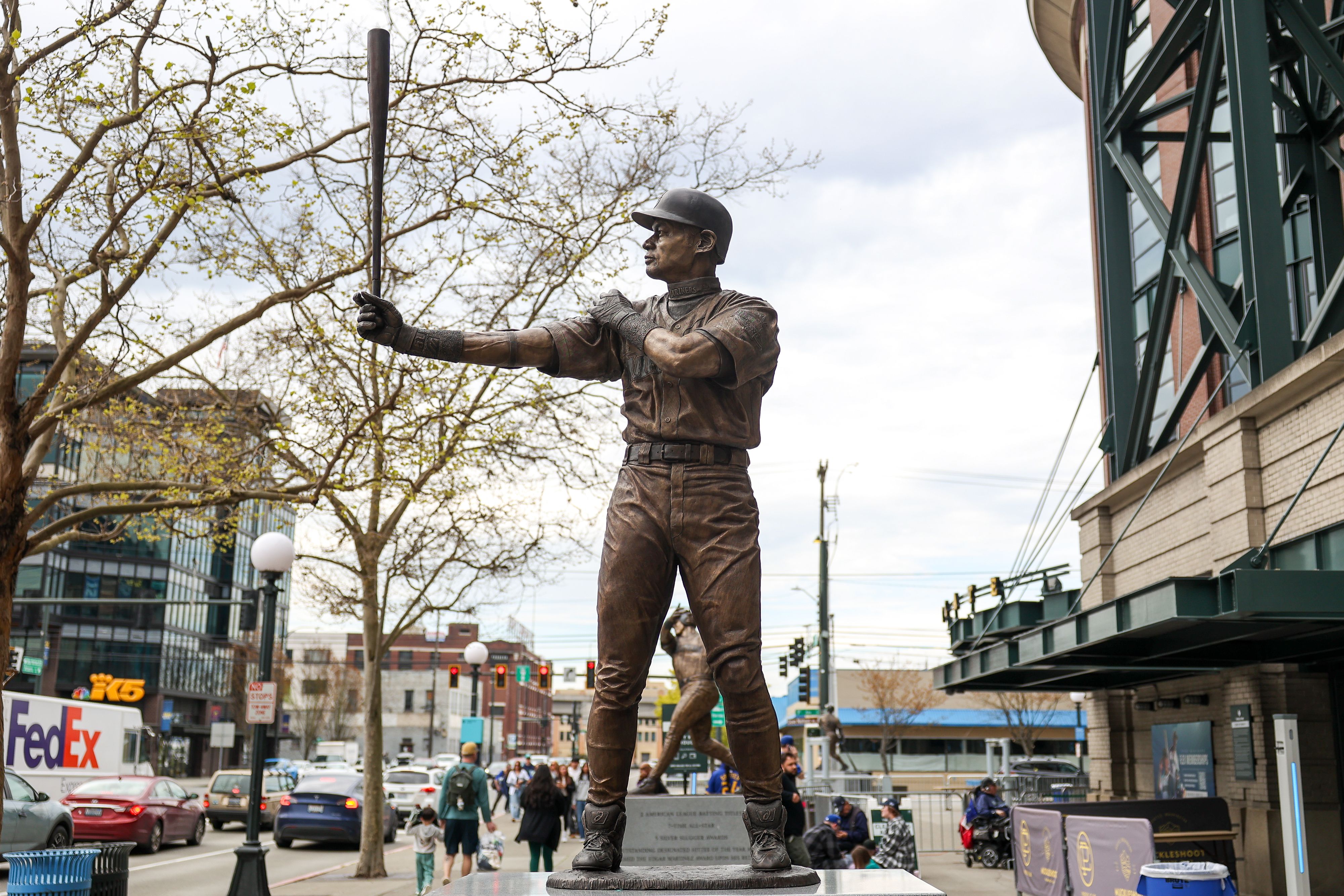 A view of a statue of Ichiro Suzuki outside T-Mobile Park, with people walking on the street and cars and traffic moving nearby.