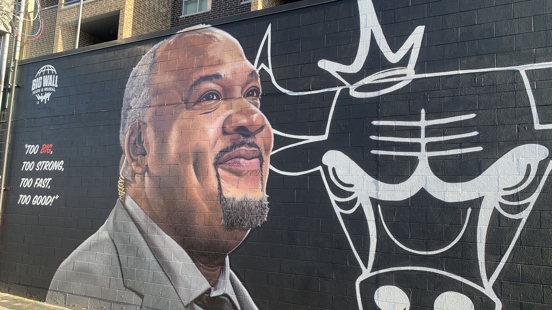 Urban mural on a dark brick wall showing a close-up portrait of a man in a suit with a gray goatee, bold white graffiti script across the right, and the text "BIG WALL SIGN & MURAL" with slogans.