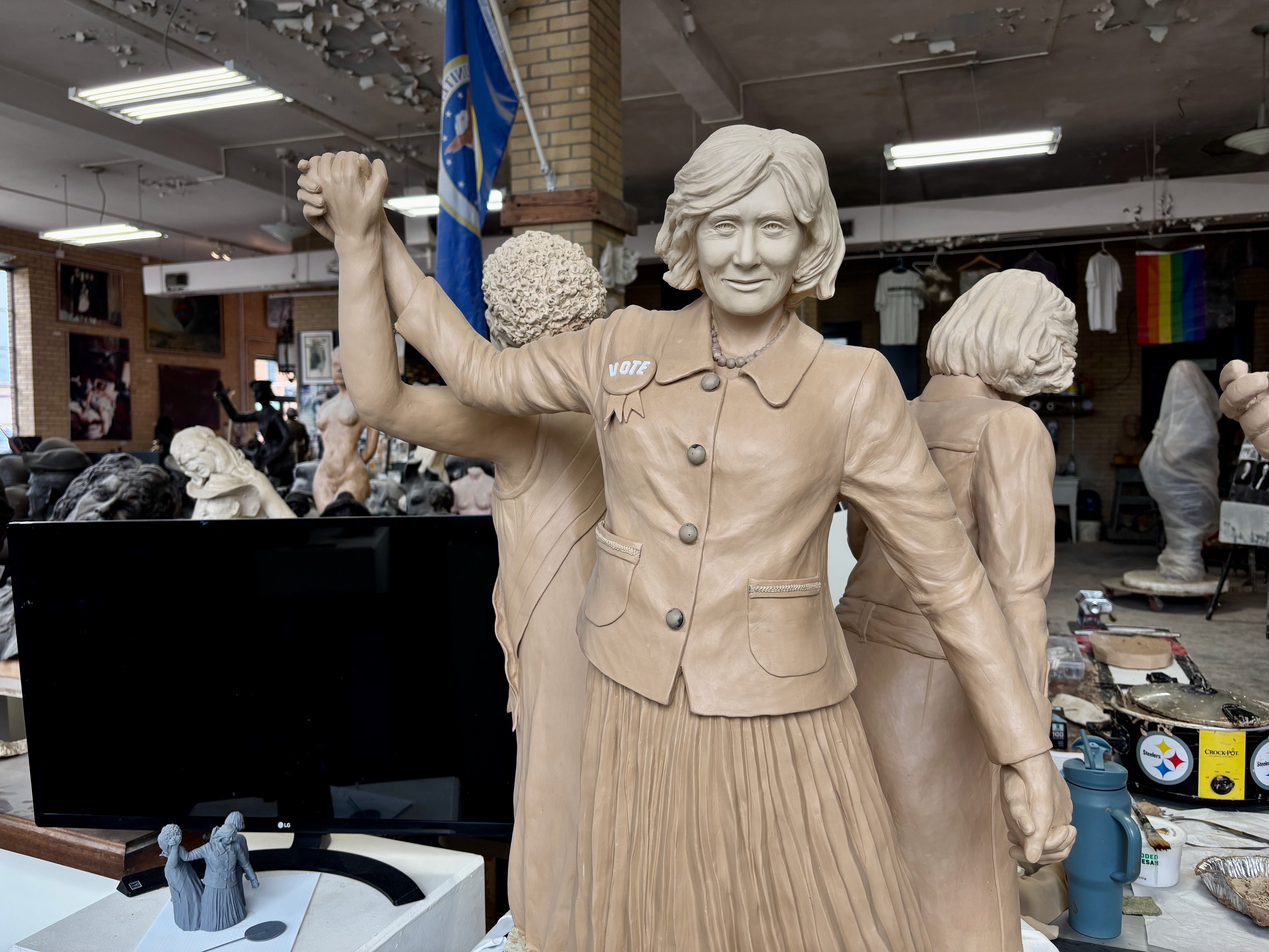 Beige clay sculpture of a woman in a blazer raising her right arm with a VOTE badge, in a busy artist studio with other statues, tools, and a rainbow flag in the background.