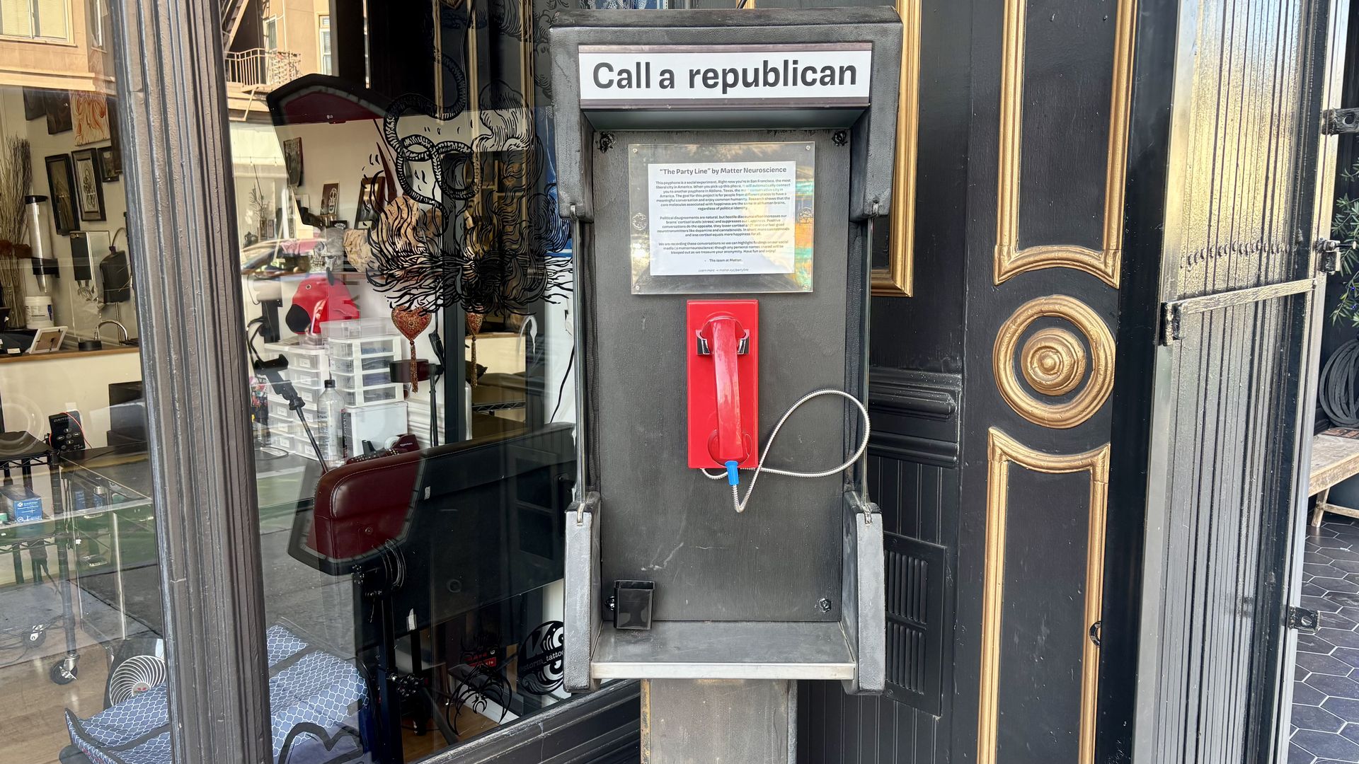 An old phone booth with a red handset and a sign above that reads "Call a republican," set against a black and gold ornate door and a shop window reflecting street and interior items.