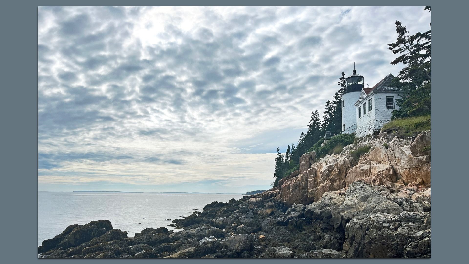 Image of Bass Harbor Head Light Station in Bass Harbor, Maine. The white lighthouse and adjoining house sit on rocky coastline with evergreen trees under a cloudy sky, overlooking calm sea with distant islands on the horizon.