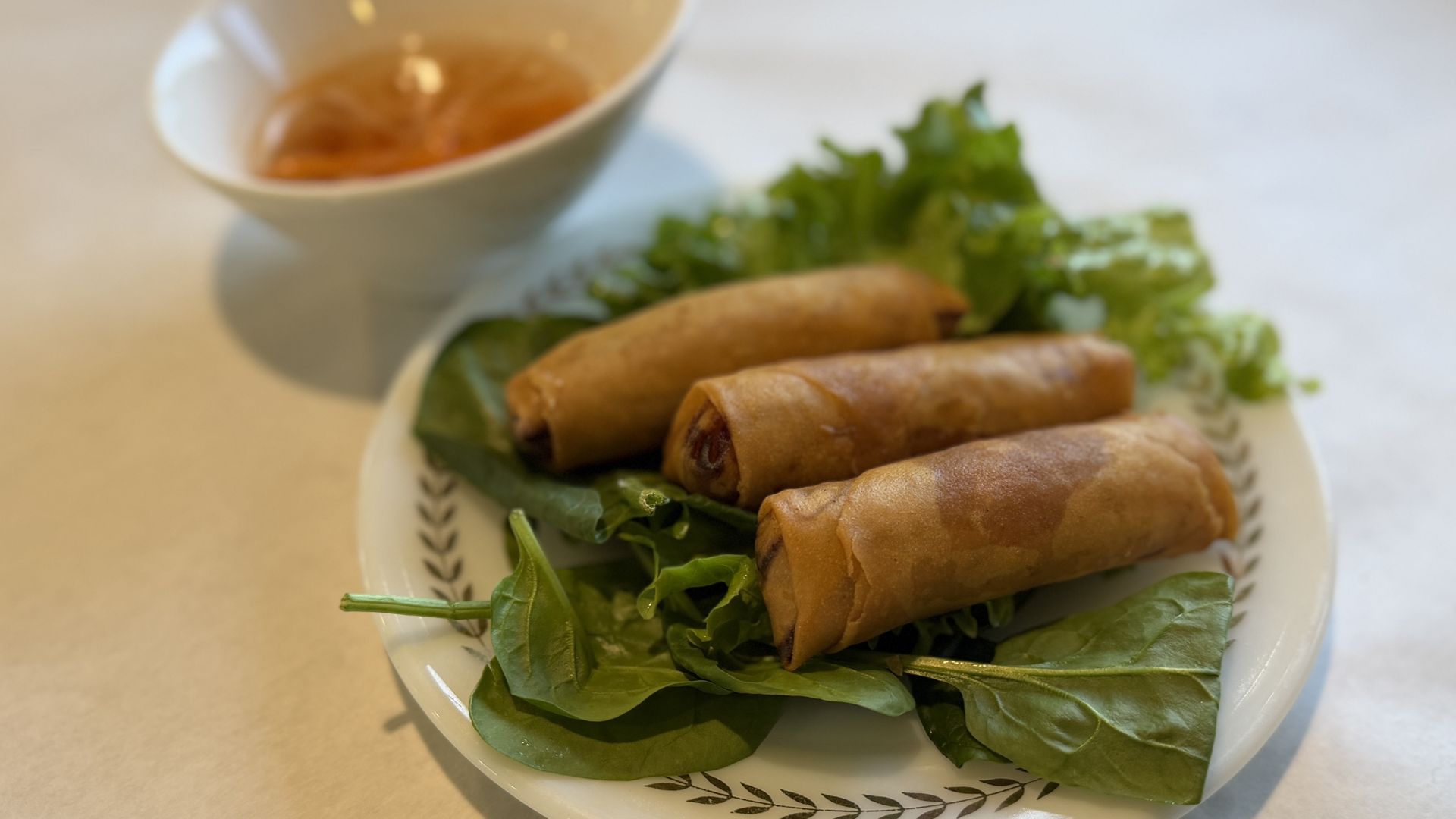 Three golden-brown fried spring rolls on a bed of fresh spinach and greens on a white plate, with a small bowl of orange dipping sauce in the blurred background.