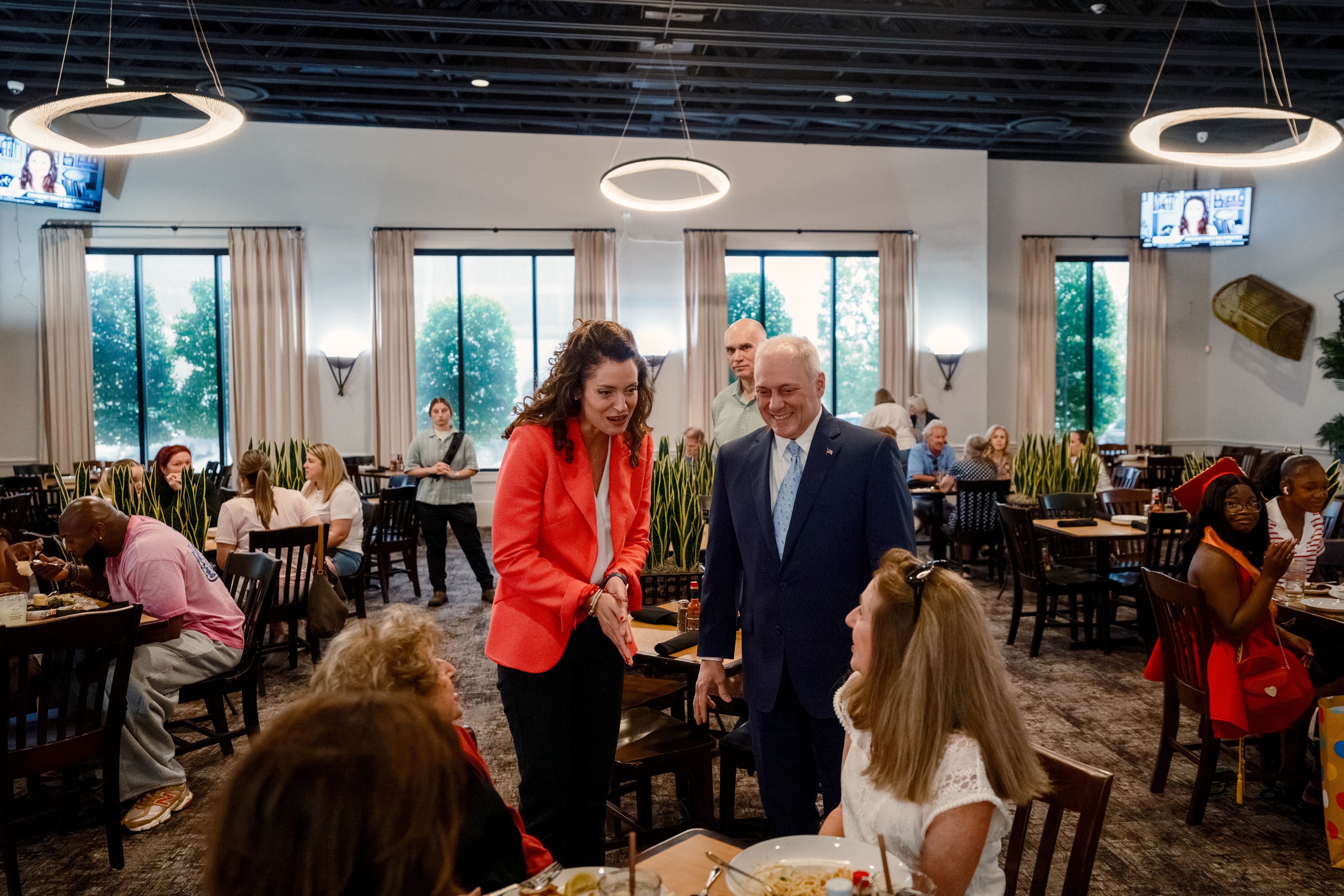 A woman in a red blazer and a man in a blue suit lean toward a table in a restaurant to talk to patrons.