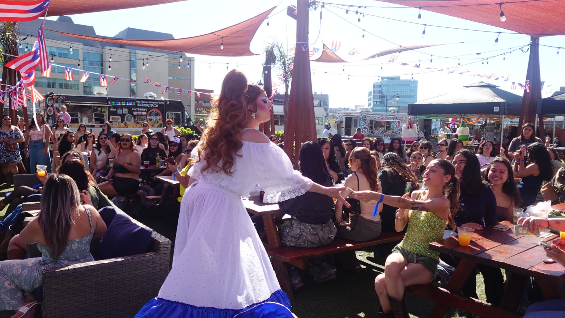 Outdoor event with many people seated at picnic tables under red shade sails and string lights. A performer in a white dress with blue trim interacts with a woman in a green top and brown boots. Puerto Rican flags hang overhead.