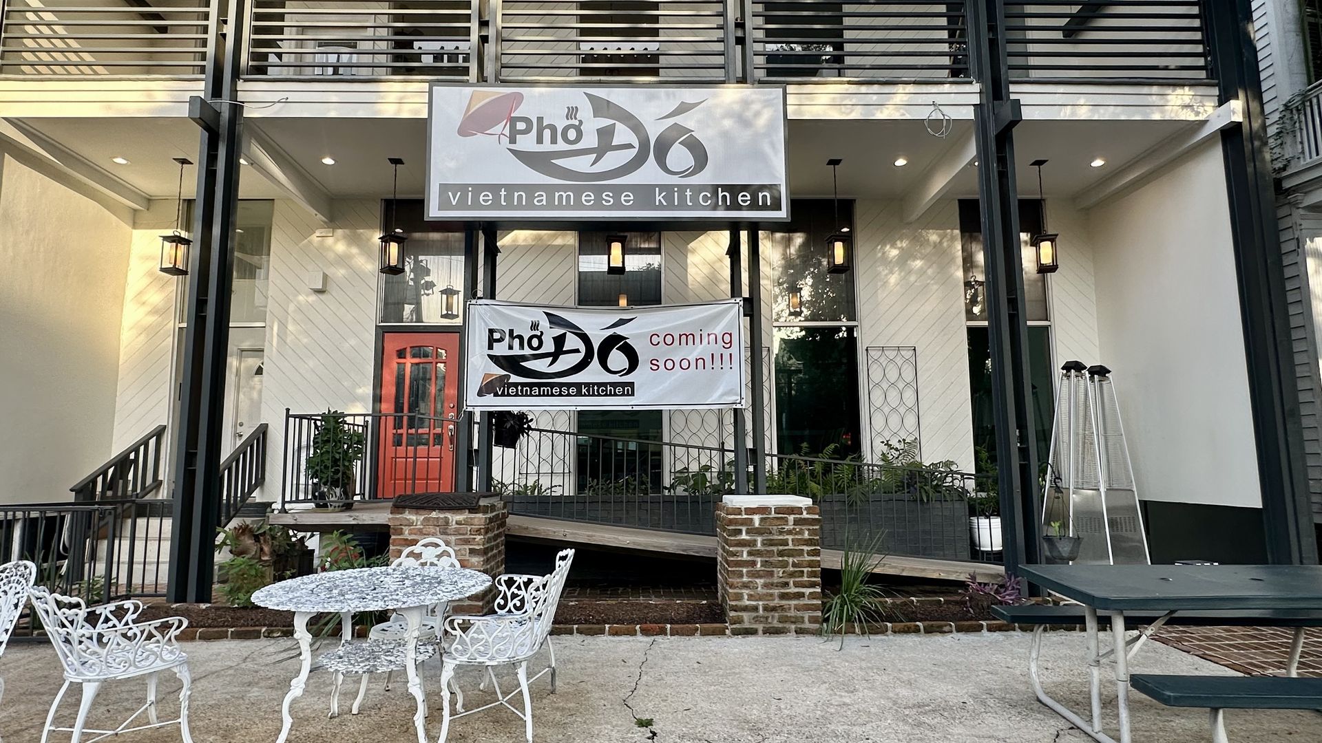 Exterior of a Vietnamese kitchen restaurant named Pho Do with signs saying "coming soon!!!" White ornate metal patio furniture and a dark green picnic table are in front.