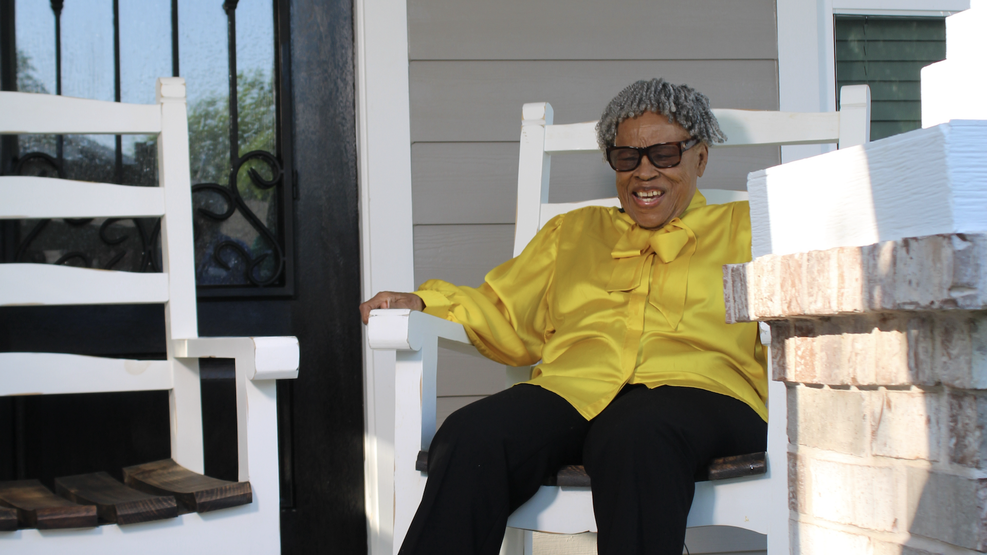 A woman wearing black and yellow sits on a rocking chair in front of a house