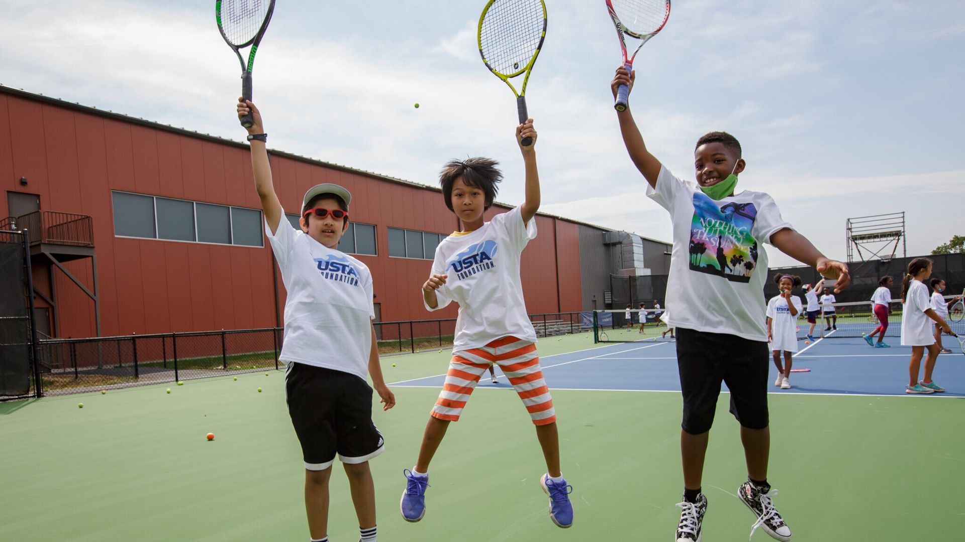 Three kids holding tennis rackets in the aid as they jump. A full tennis court of kids behind them.