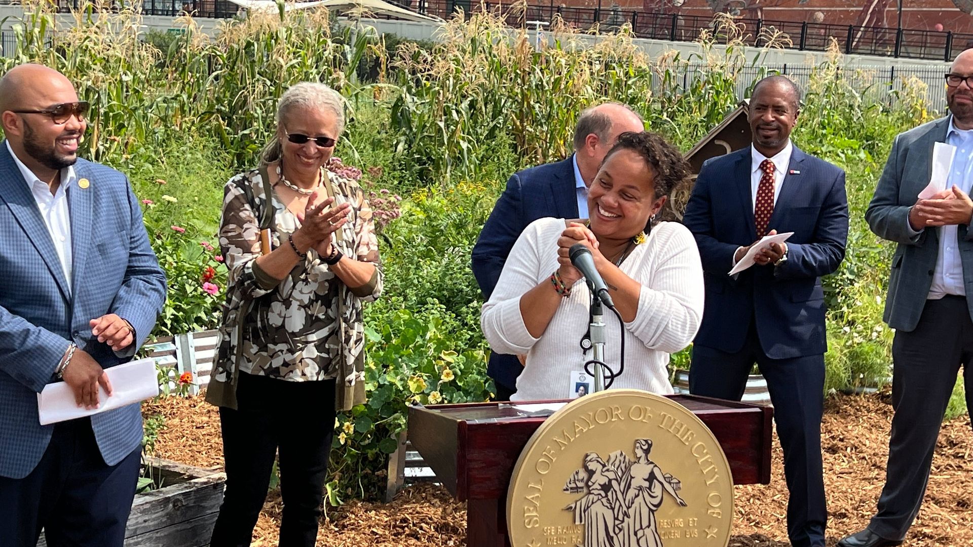 New director of agriculture Tepfirah Rushdan stands at a podium smiling in front of other officials and a garden, with brick buildings in the background.