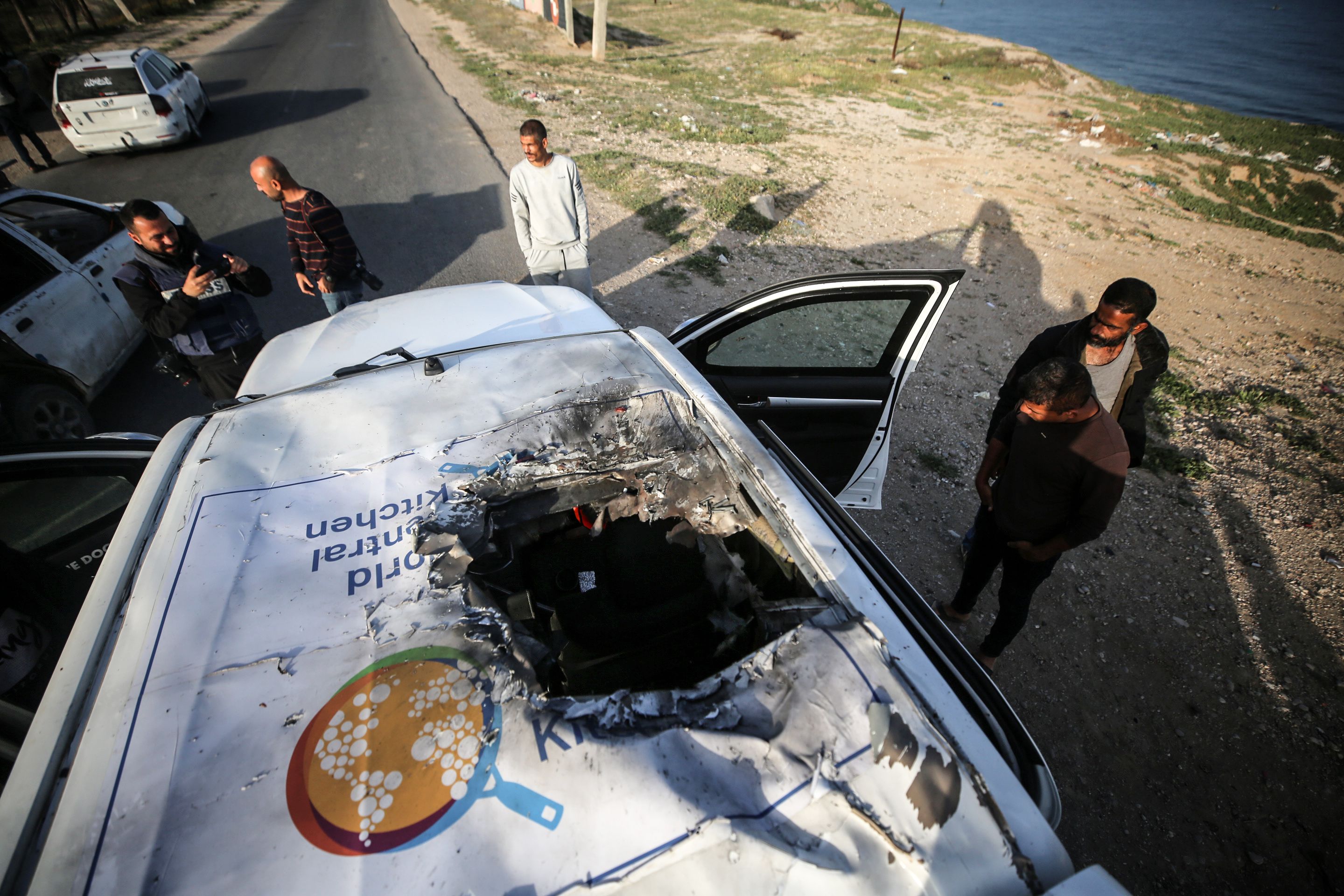 Palestinians standing next to a vehicle with a World Central Kitchen logo and a bombed out roof.