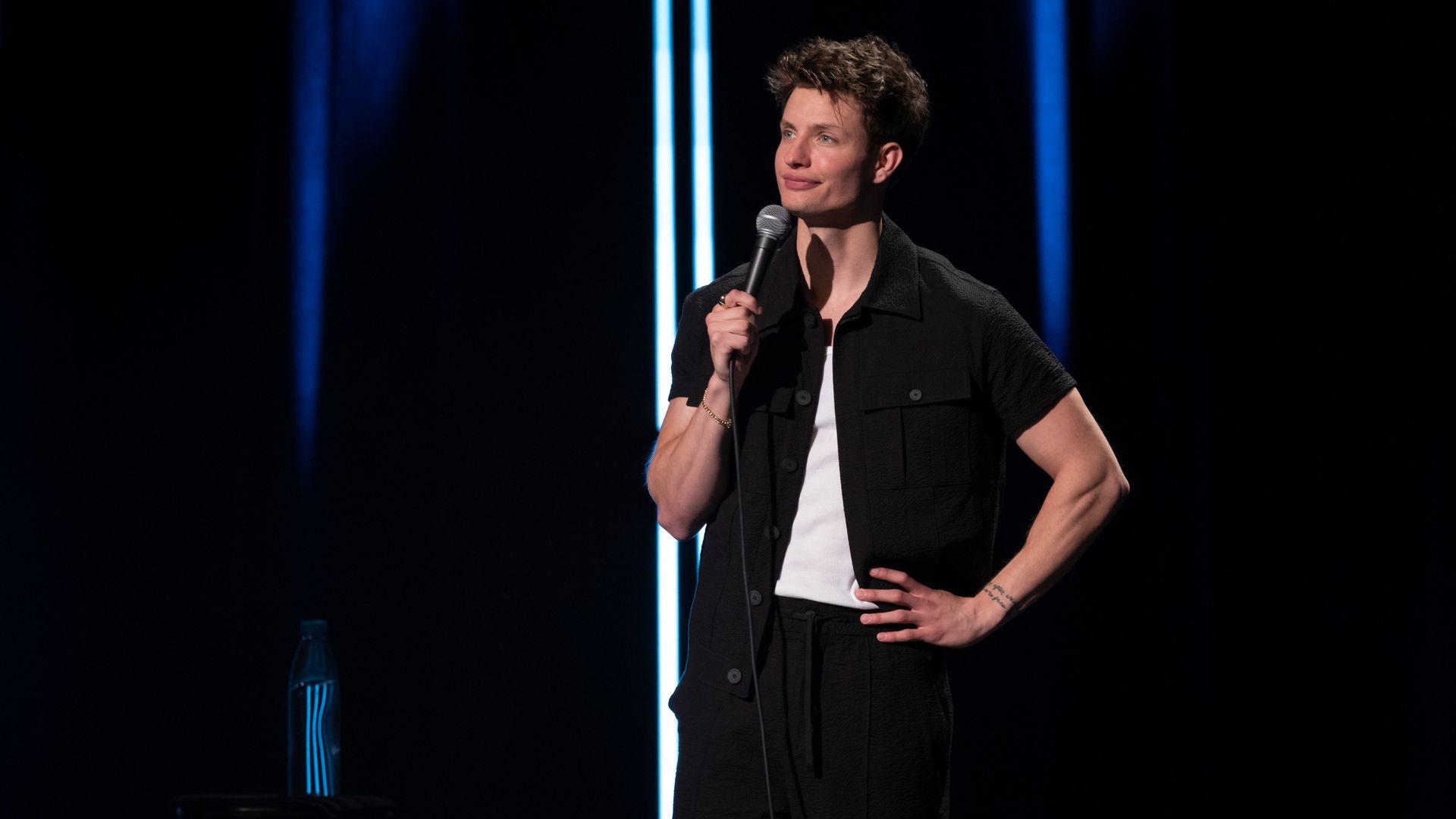 Comedian on a dark stage holds a microphone, wearing a black short-sleeve shirt over a white tee; blue vertical lights in the background, with a water bottle on a stool to the left.