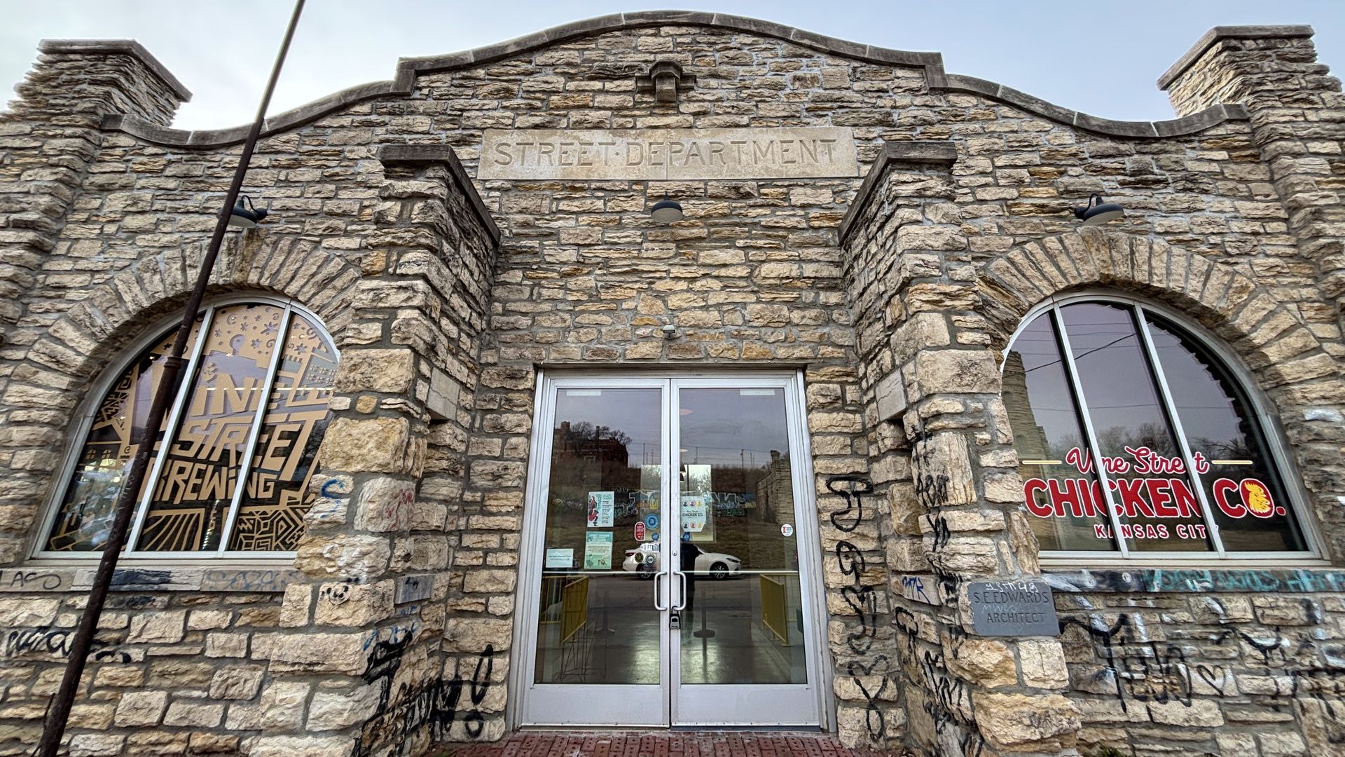 Stone building with arched windows and graffiti, labeled "Street Department". Left window shows "Indie Street Brewing" logo, right window shows "Vine Street Chicken Co. Kansas City" sign.