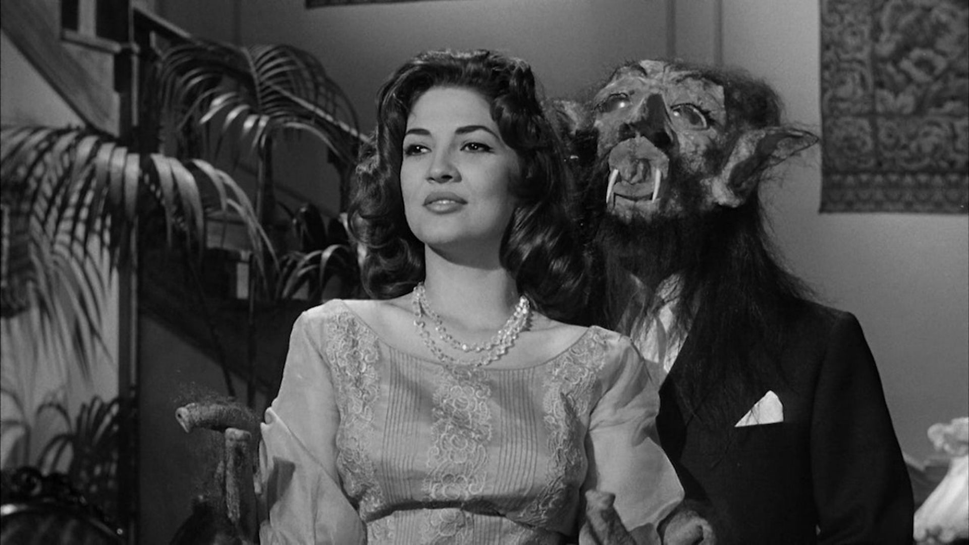 Black and white image of a woman in a lace dress and pearl necklace sitting in front of a man wearing a monster mask with fangs and long hair in a suit.