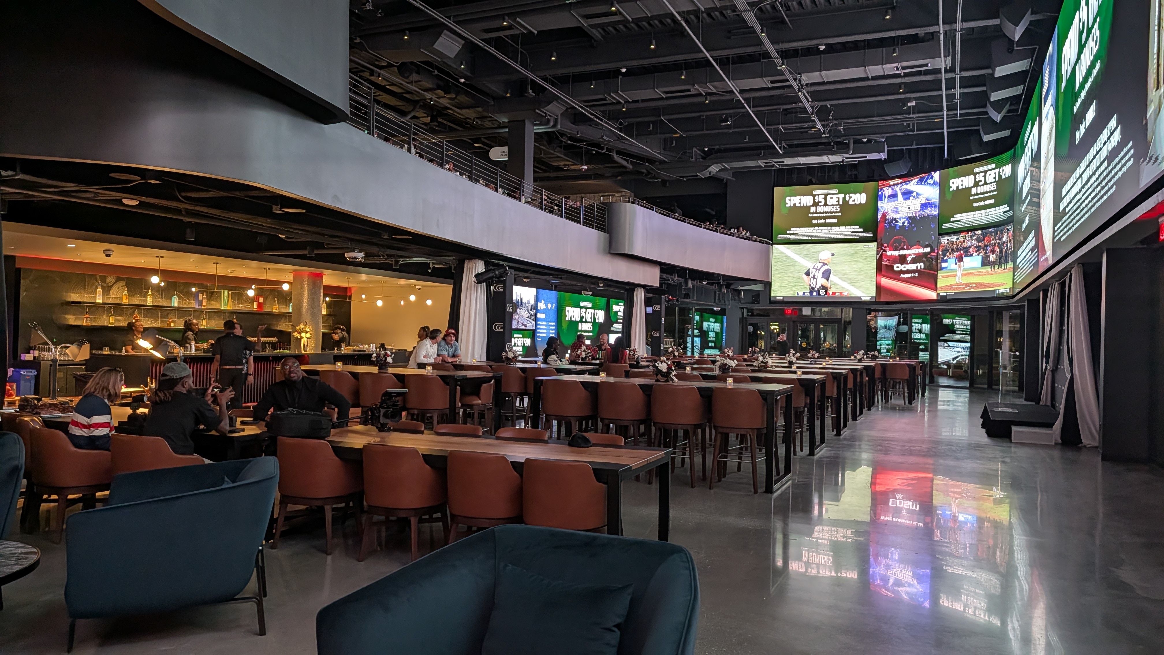 Interior of a modern sports bar with numerous large stadium screens, a long bar on the left, rows of brown chairs, lounge seating in foreground, and a dark ceiling with exposed wiring.