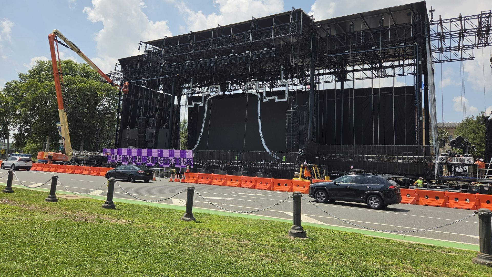 Outdoor concert stage under construction with a large black metal rig, scaffolding and lighting. A yellow boom lift on the left, orange barriers along road, green grass, and a blue sky with clouds.