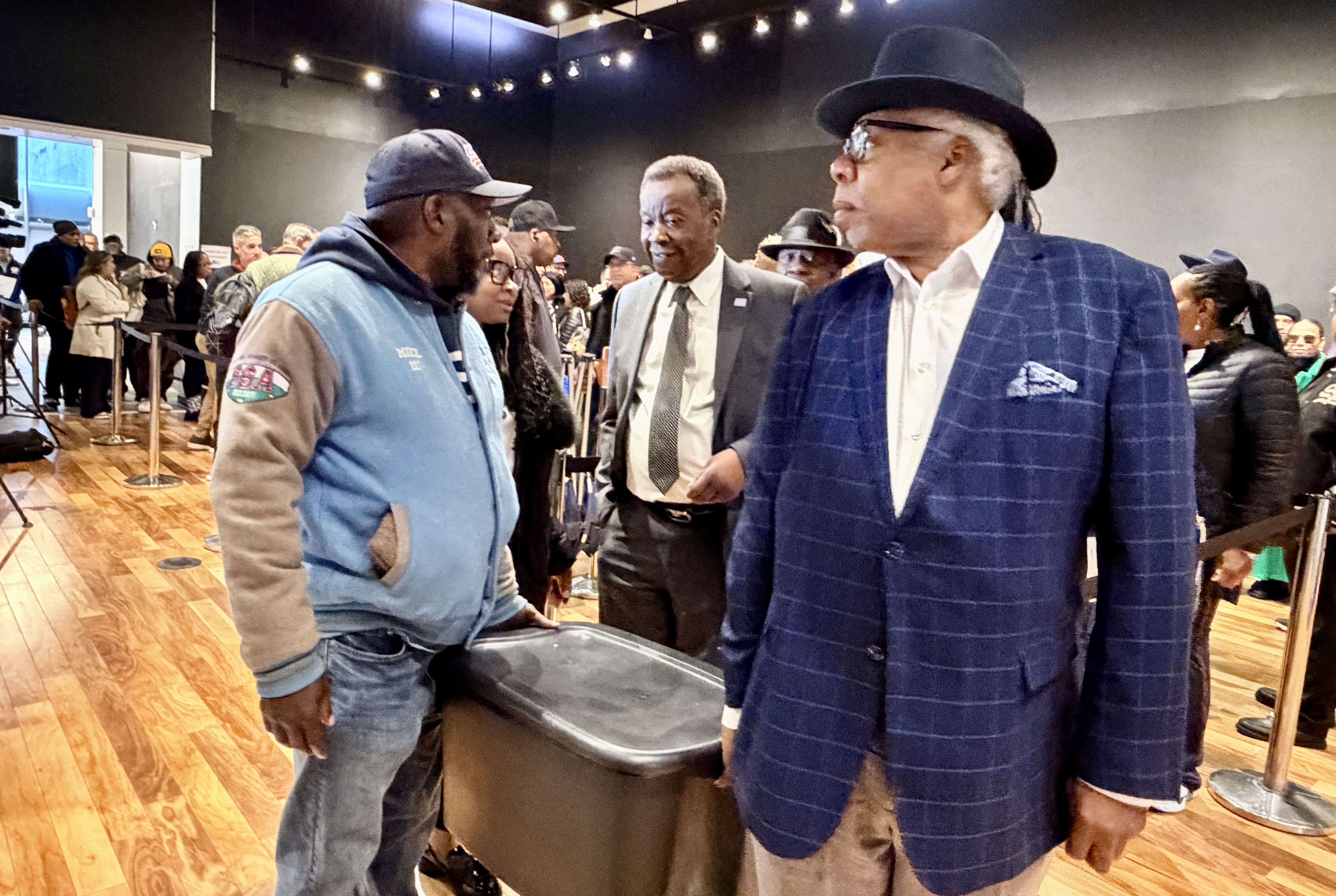 Two men converse at an indoor event: one in a blue plaid suit and hat on the right, the other in a blue jacket and cap on the left, with a crowd and wooden floor behind them.