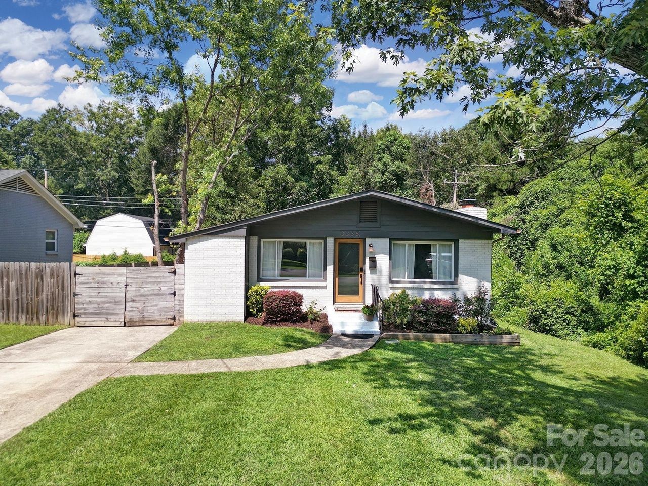 House for sale charlotte. Small single-story white brick house with a dark gray roof, wooden front door, two windows, and a curved concrete path to the steps. Green lawn, shrubs, driveway on the left, and trees in the yard.