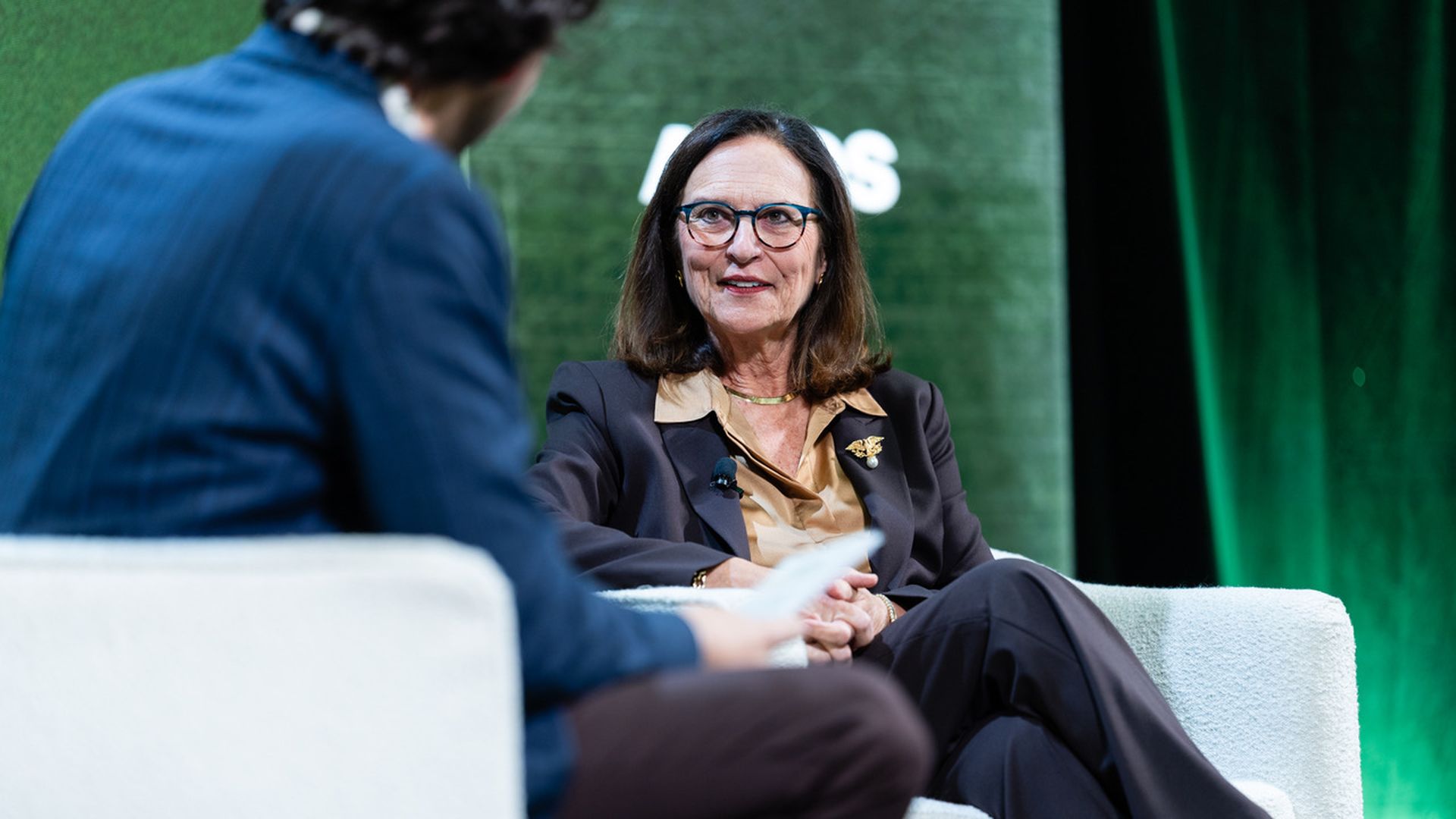 A woman in a dark brown suit and glasses sits on a white chair talking to a man in a blue jacket, with a green backdrop behind them at an Axios event.