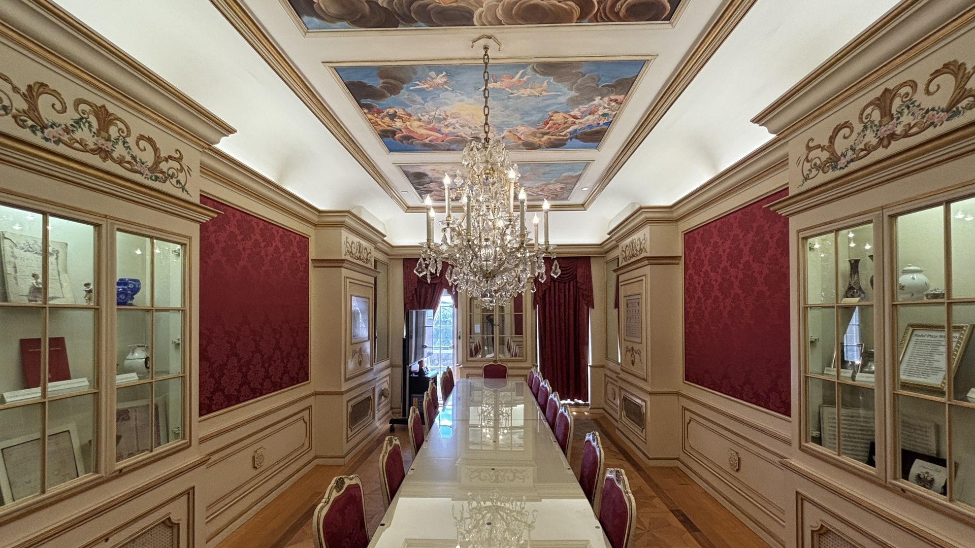 Luxurious antique gallery/dining room with a long glass table, red damask walls, gold trim, and a crystal chandelier. Ceiling fresco panels featuring cherubs; glass display cabinets line both sides.