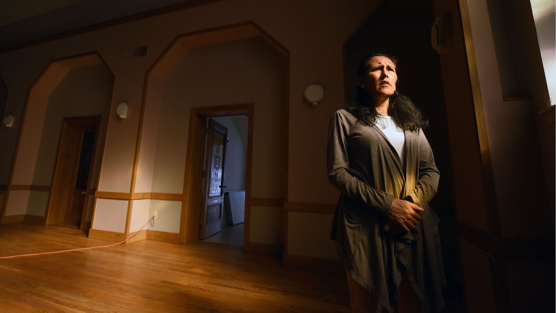 In this image, a woman stands facing an unseen window as light falls on her face.