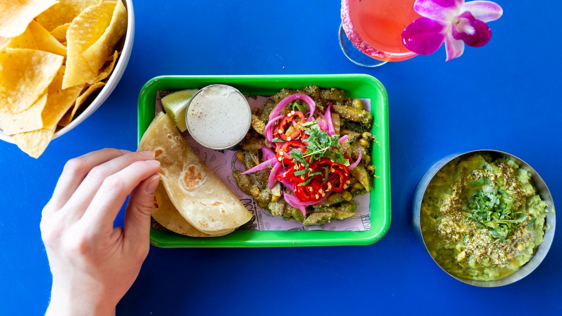 A hand reaches for a flour tortilla and taco fillings on a rectangular green plate. The blue table is also set with tortilla chips, guacamole and a pink cocktail.