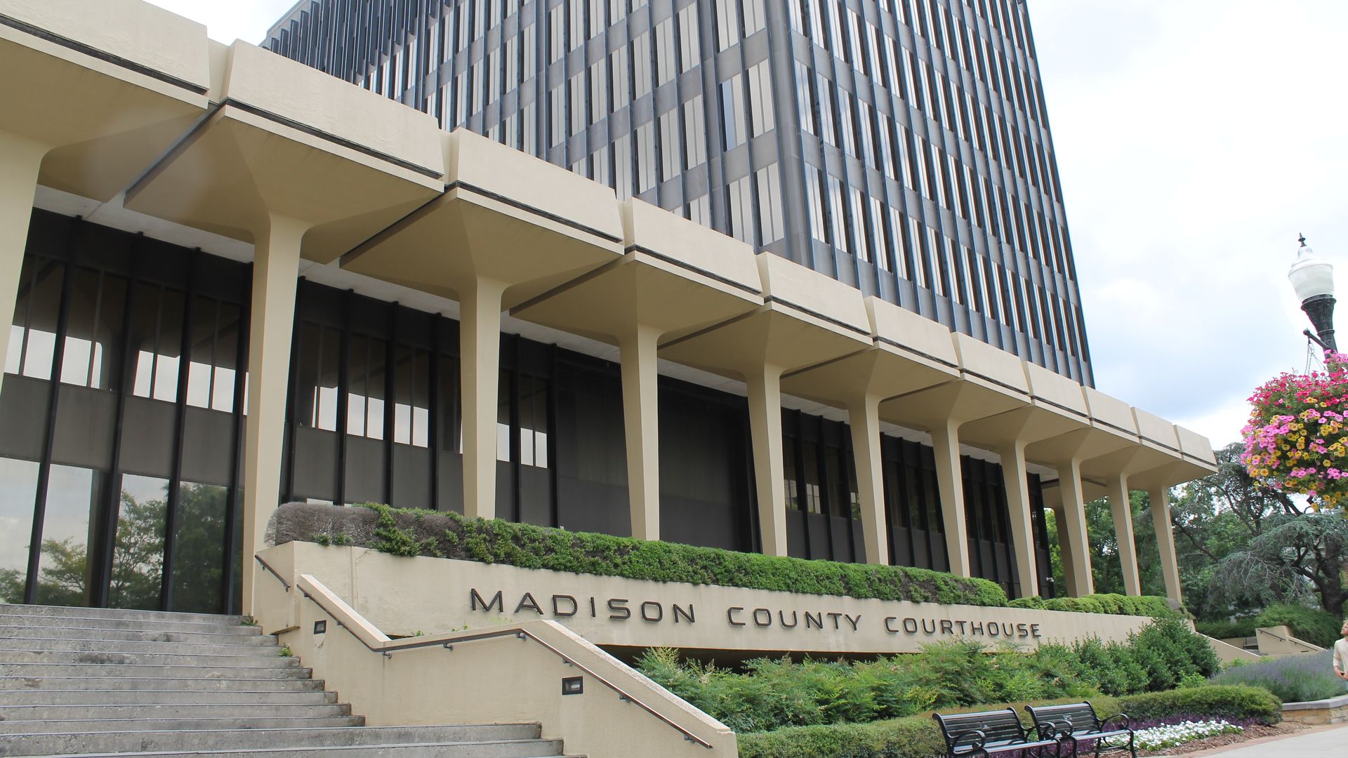 Front view of a modern courthouse with beige columns and a tall glass facade. Stairs lead to the entrance; manicured shrubs and flowers line the walkway beside the Madison County Courthouse sign.