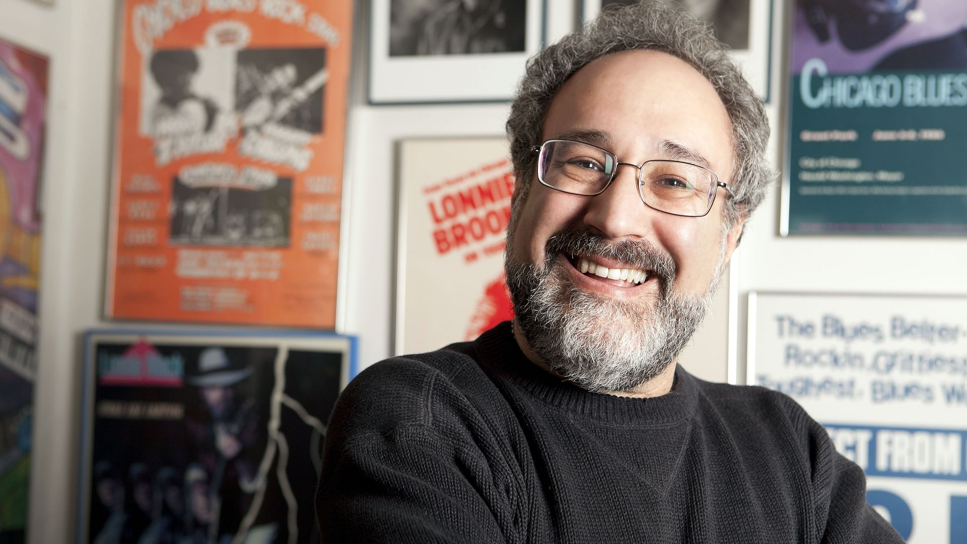 A smiling man with gray hair and a beard, wearing glasses and a dark sweater, posing in a room with colorful posters on the wall.