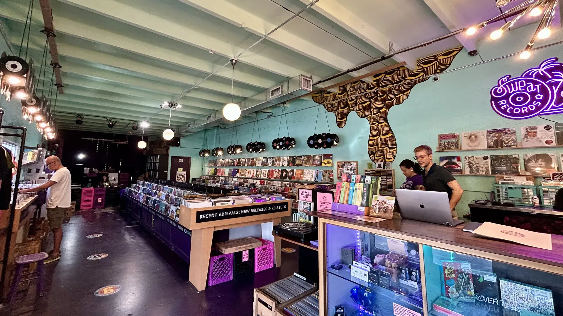 Inside a turquoise-walled record shop with vinyl racks, hanging globe lights, and a neon "Sweet Records" sign. 