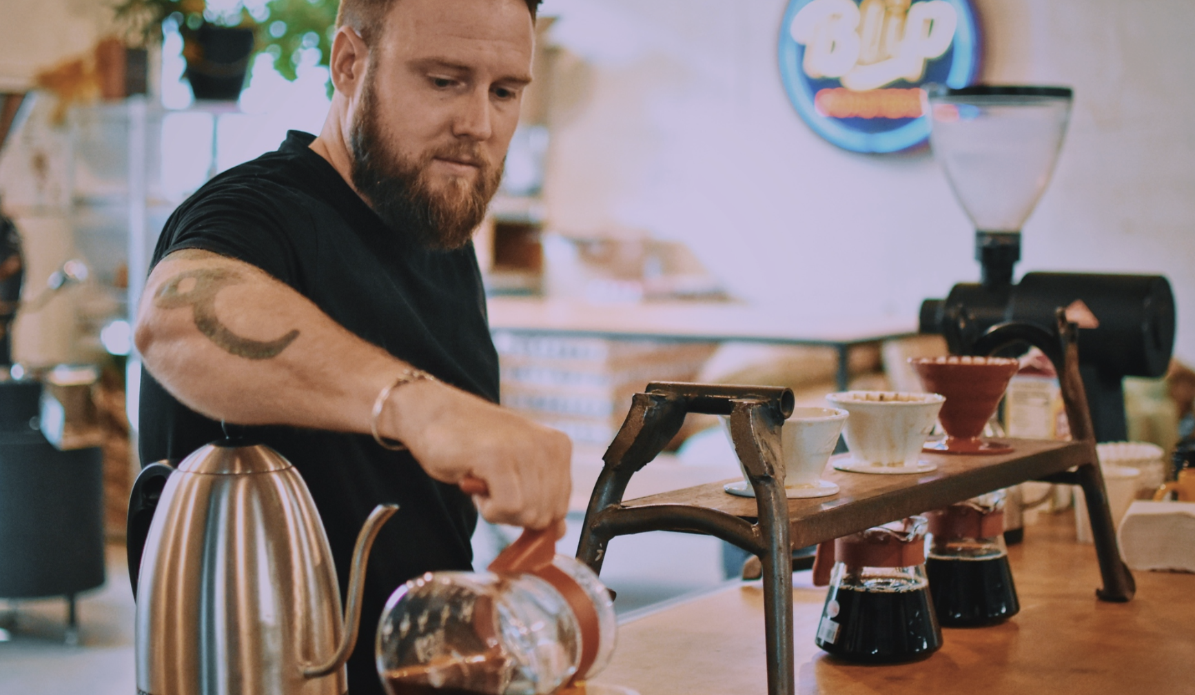 A bearded man in a black shirt with a tattoo on his arm pours coffee using a glass pot at a wooden stand holding three coffee drippers in a cozy, softly lit cafe.