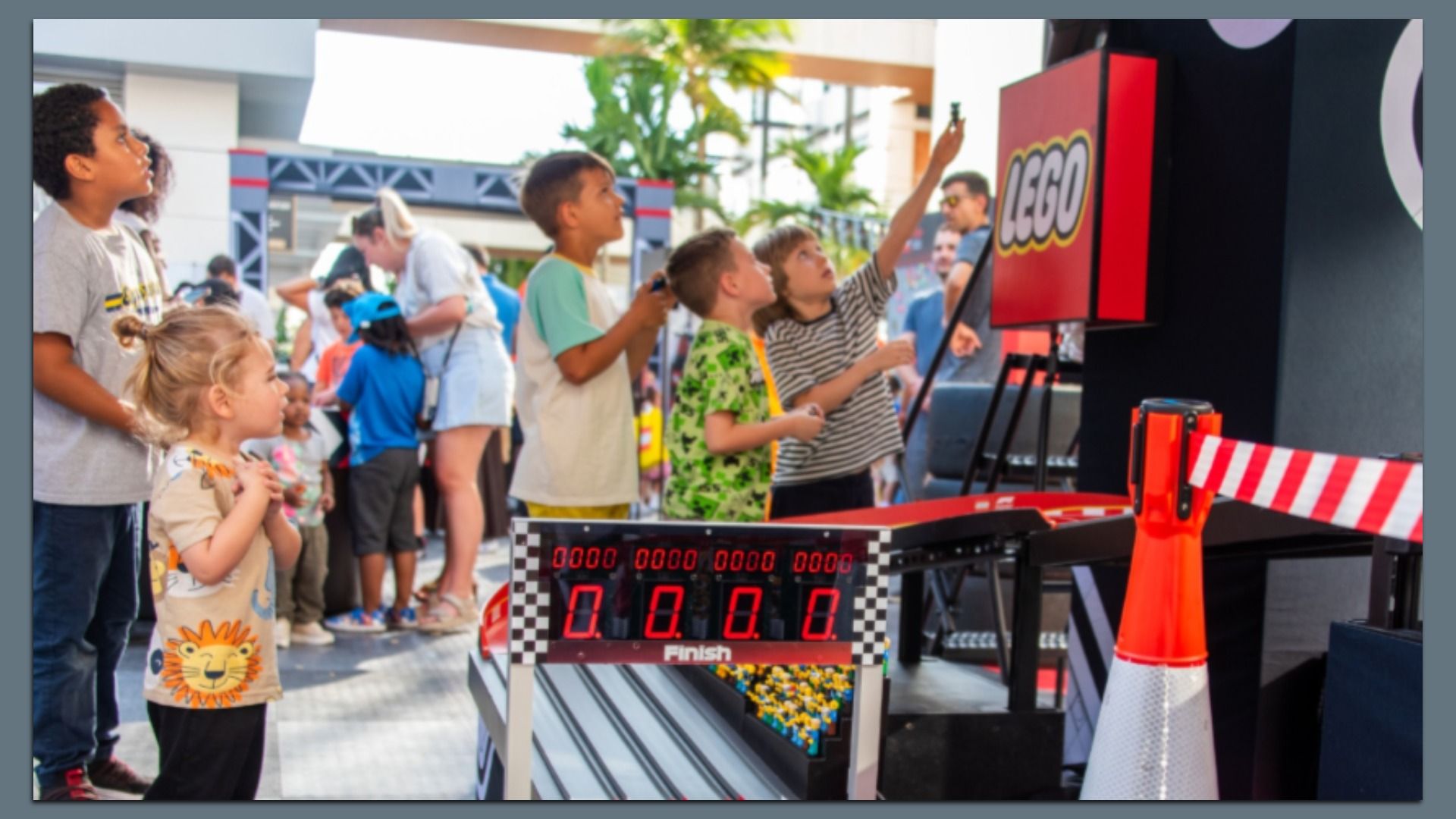 Children crowd around a LEGO interactive race display outdoors; a boy points at a red LEGO screen while a digital finish board shows zeroes and a checkered track, with palm trees in the background.