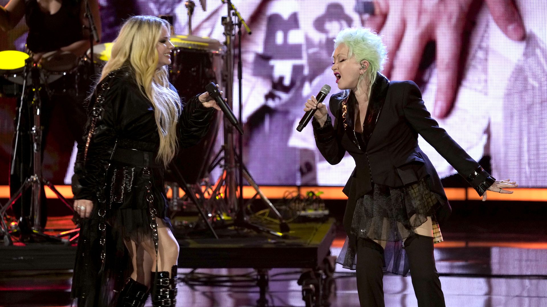 Two female singers performing on stage with microphones; the one on the left has long blonde hair and a black outfit, the one on the right has short mint green hair and a black blazer. Drums in background.
