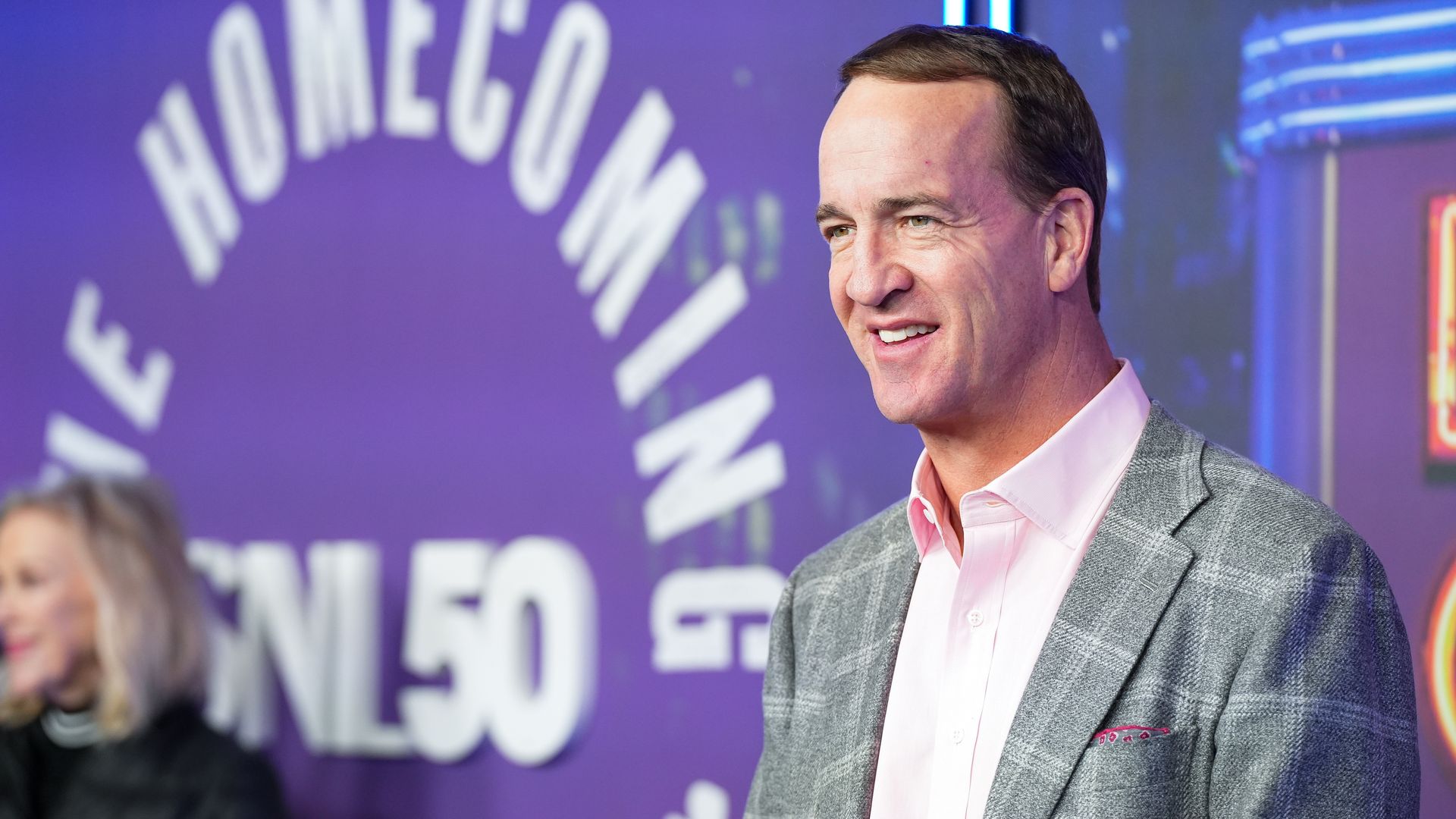 A man in a gray blazer and pink shirt looks on. 