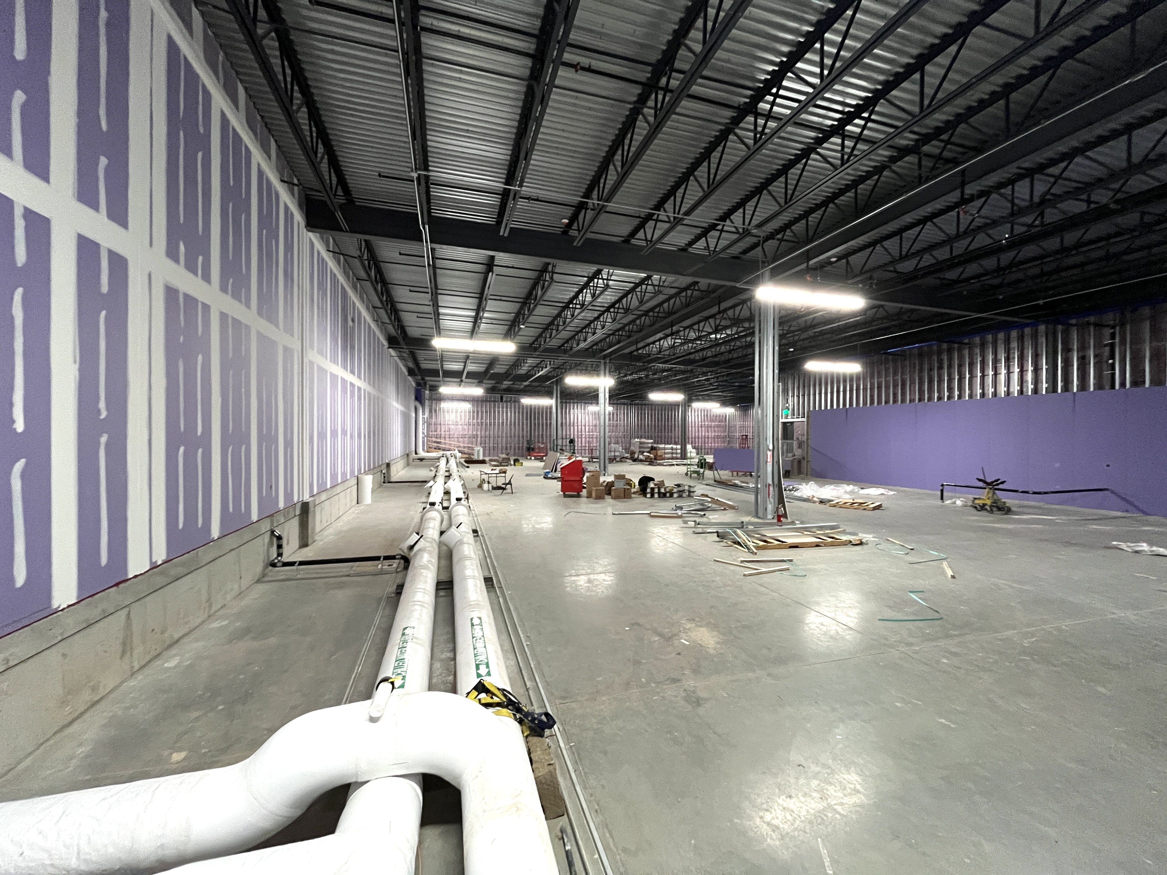 Large industrial space under construction with exposed steel ceiling, purple and white walls, large white pipes on the left, scattered tools and materials on concrete floor, bright overhead lights.