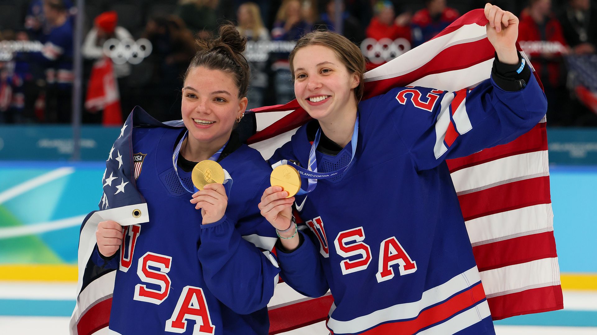 Cayla Barnes and Hannah Bilka, wearing blue USA jerseys hold gold medals and an American flag behind them, celebrate on the ice rink with a blurred audience in the background.