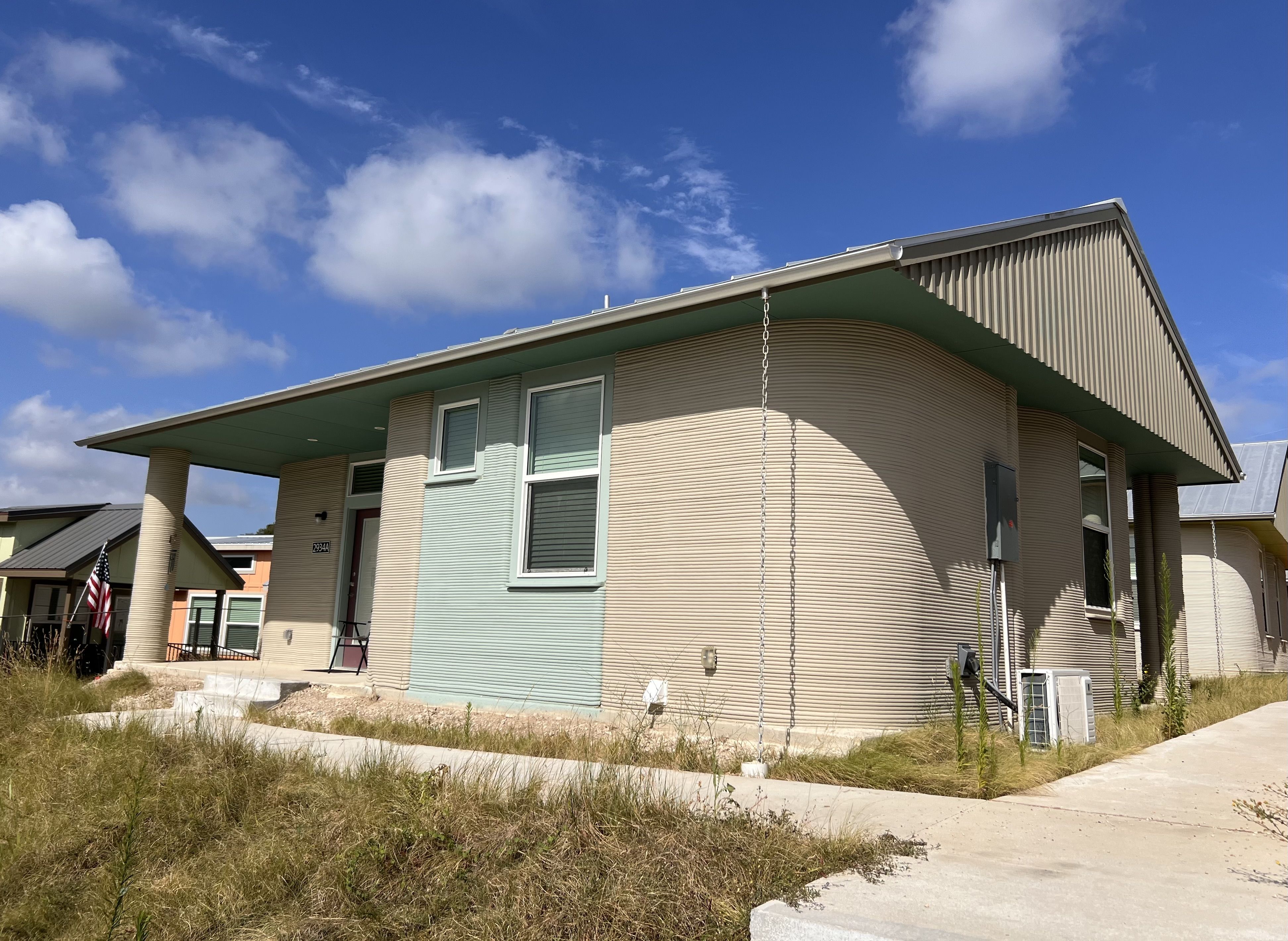 Beige house with teal trim and curved walls, under a bright blue sky with clouds. A hanging chain from the roof overhang, large windows, dry grass foreground, and a small American flag on the left.