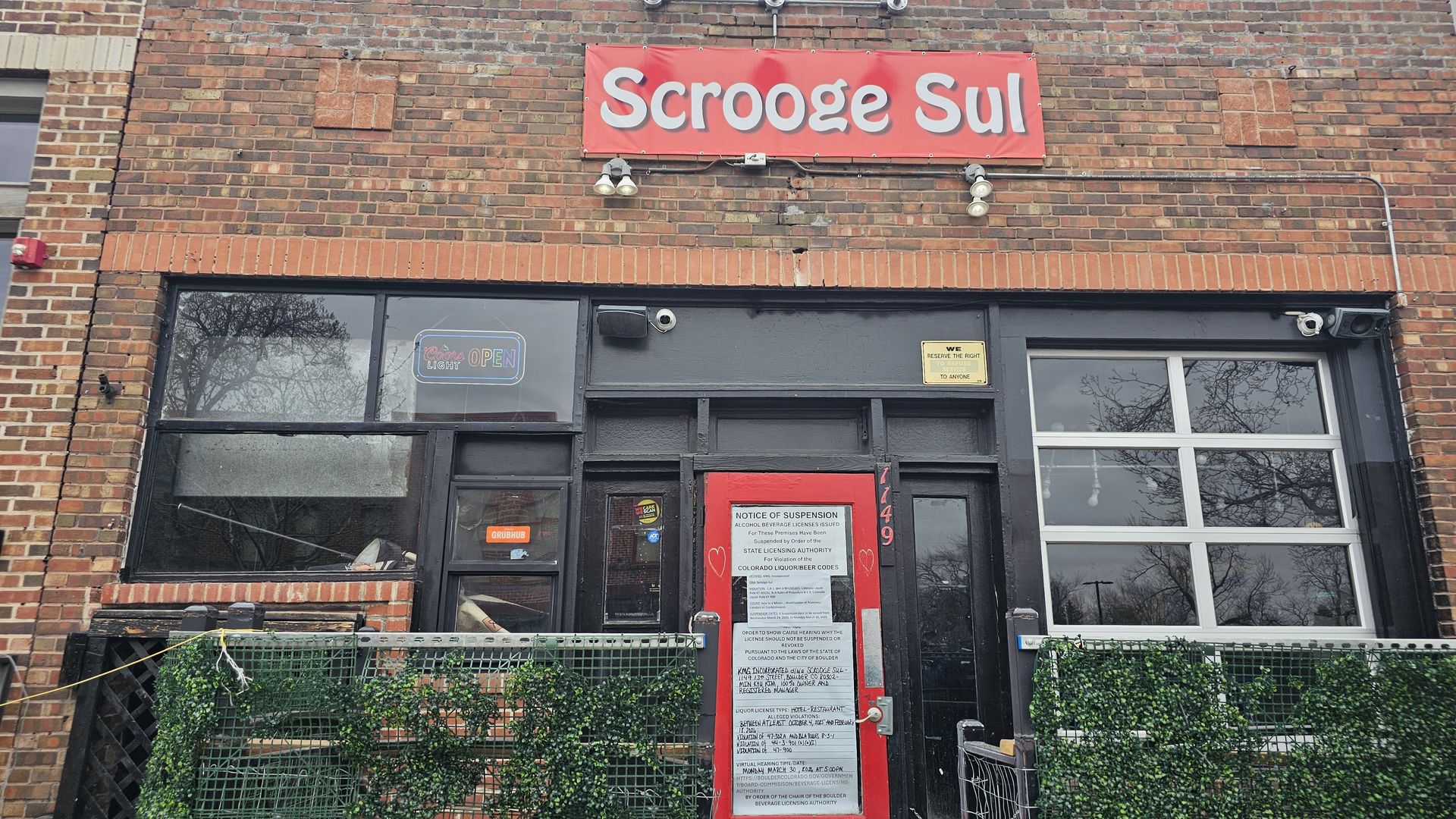 Brick storefront with a red sign reading Scrooge Sul above a dark door. Large front windows, and a green fence with ivy; a neon OPEN sign is visible in the left window, and a posted notice on red door.