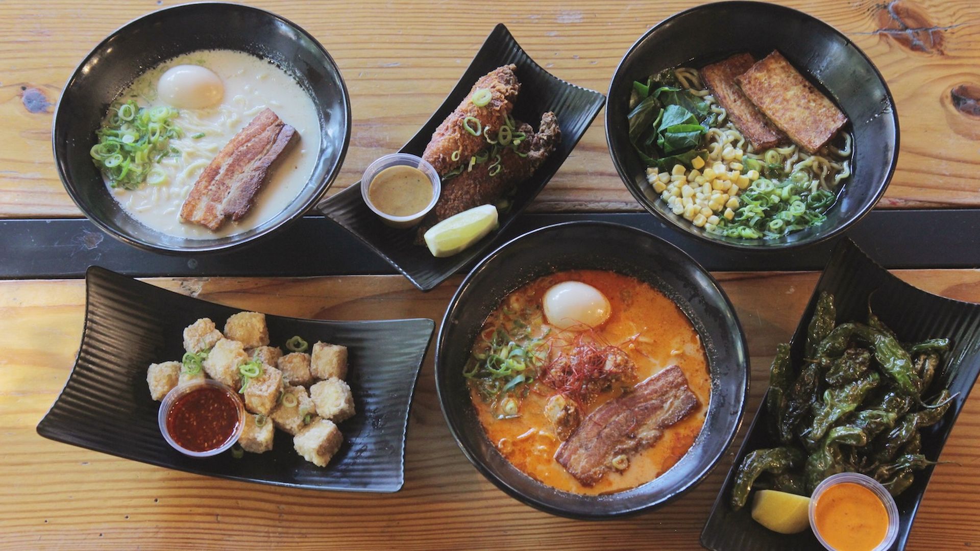 A spread of Japanese-inspired dishes including two ramen bowls with pork, egg, and green onions, a vegan ramen with tofu, fried chicken, crispy tofu bites, shishito peppers, and dipping sauces.