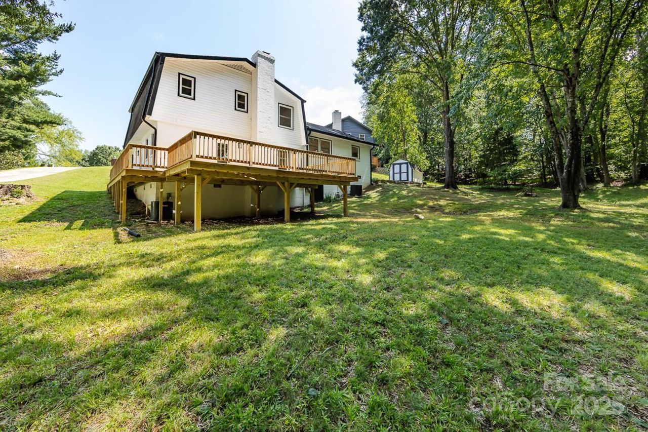White two-story house with black roof and chimney, wooden deck, large green lawn, and surrounding tall trees under a clear blue sky on a sunny day.