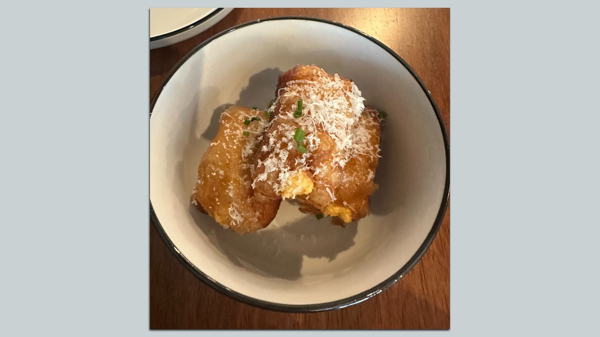 Two golden fried pastry bites in a white bowl with a black rim, topped with grated white cheese and chopped green onions, on a wooden table.