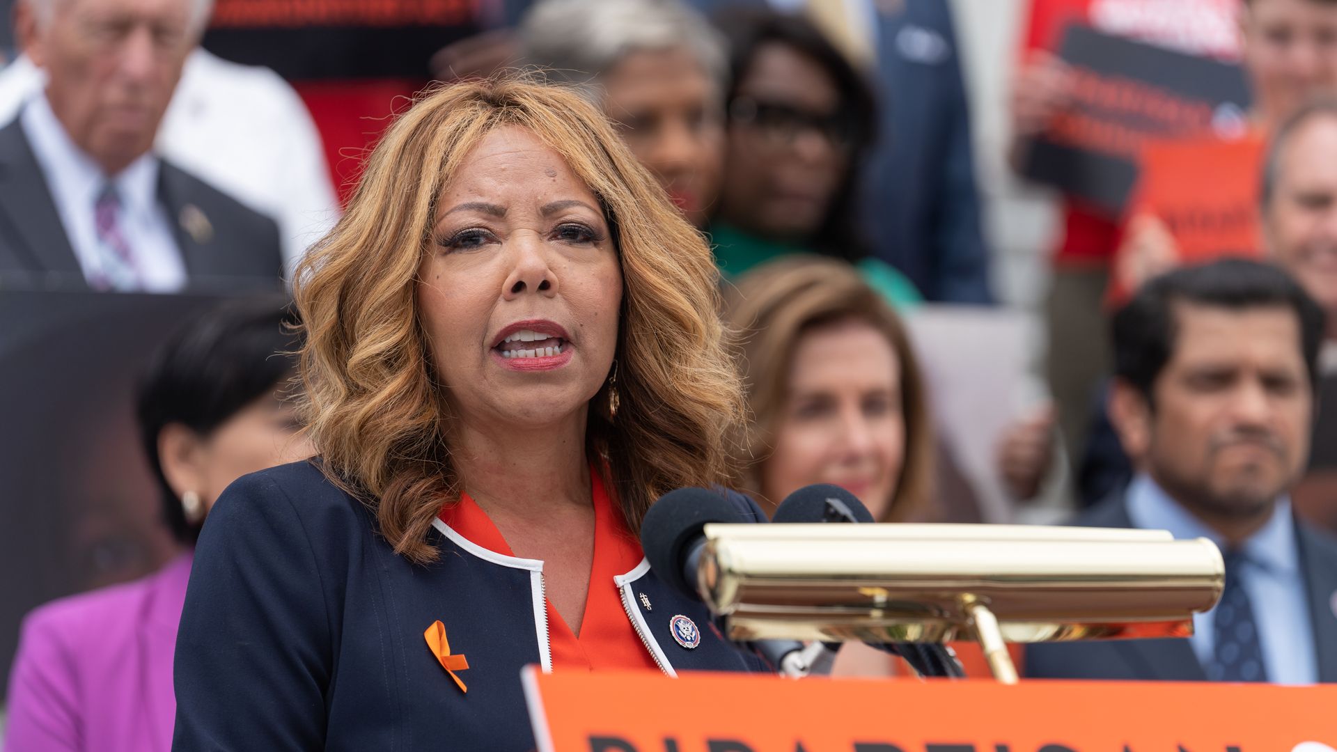 A woman lawmaker standing at a lectern bearing an orange sign with the word "bipartisan" speaks surrounded by lawmakers